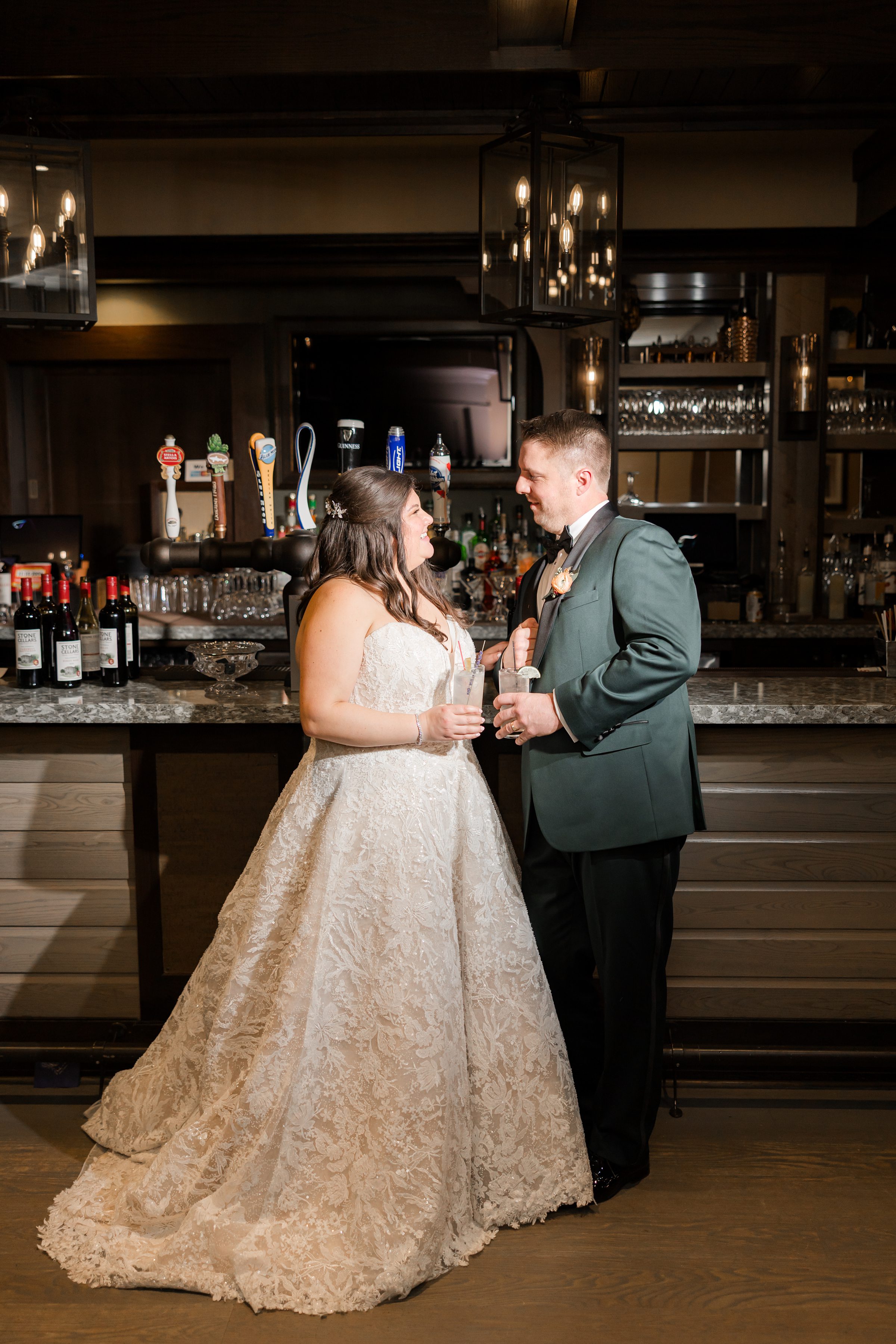 Bride and groom stand at a bar, smiling at each other while holding drinks under warm, dim lighting