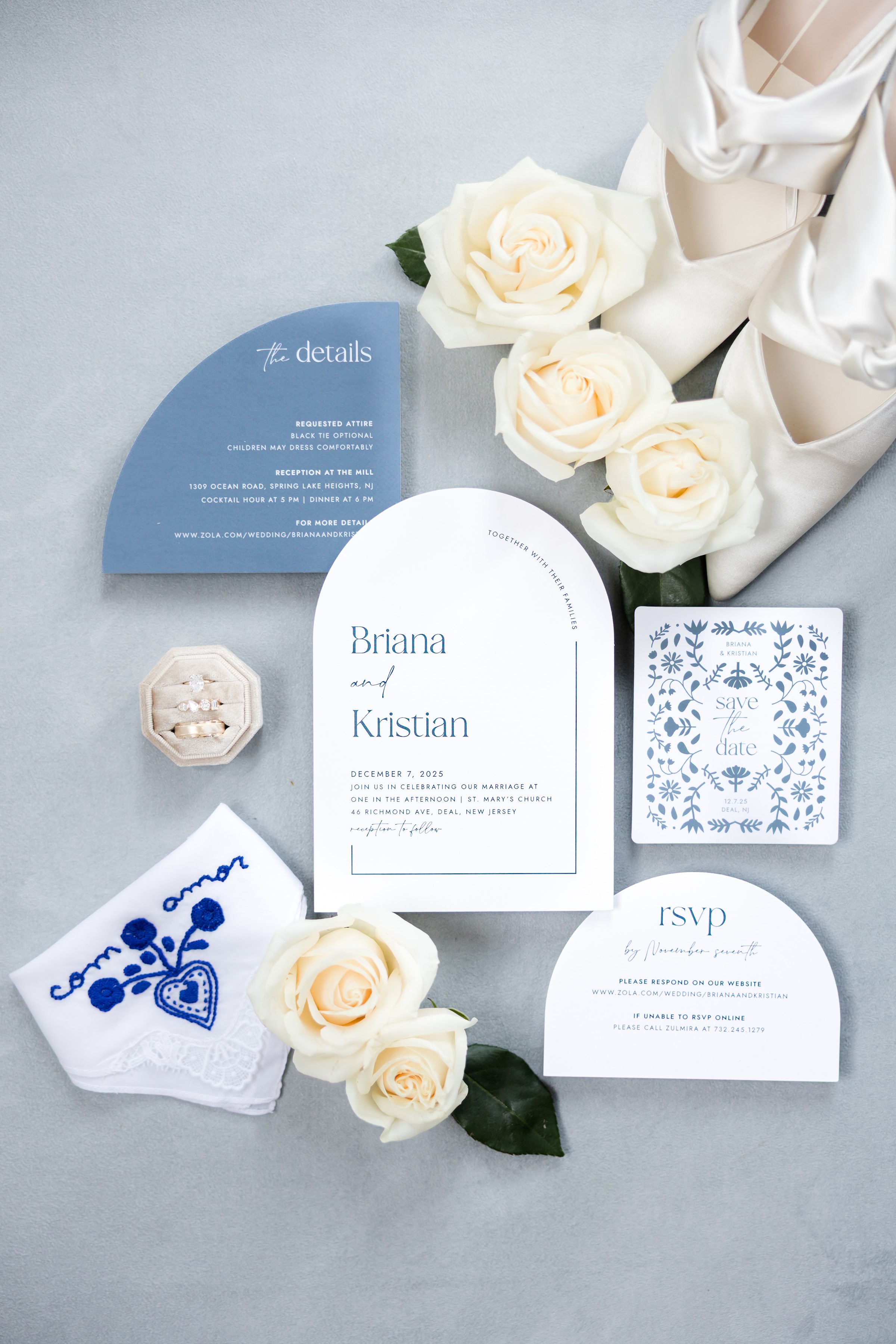 Wedding stationery suite featuring invitations, rings, bridal shoes, an embroidered handkerchief, and white roses 