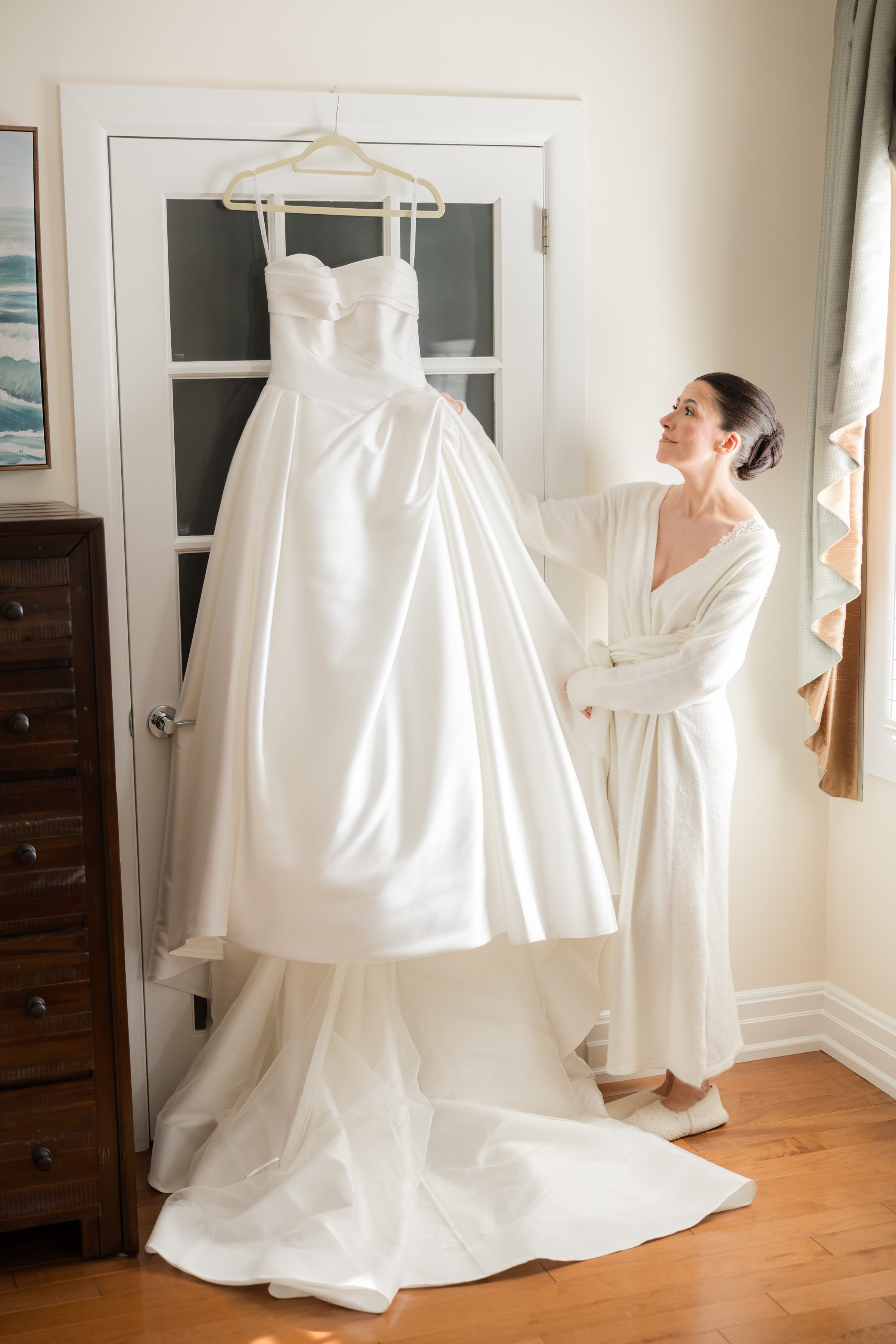 Bride in a robe looking at her white wedding dress hanging on a door.
