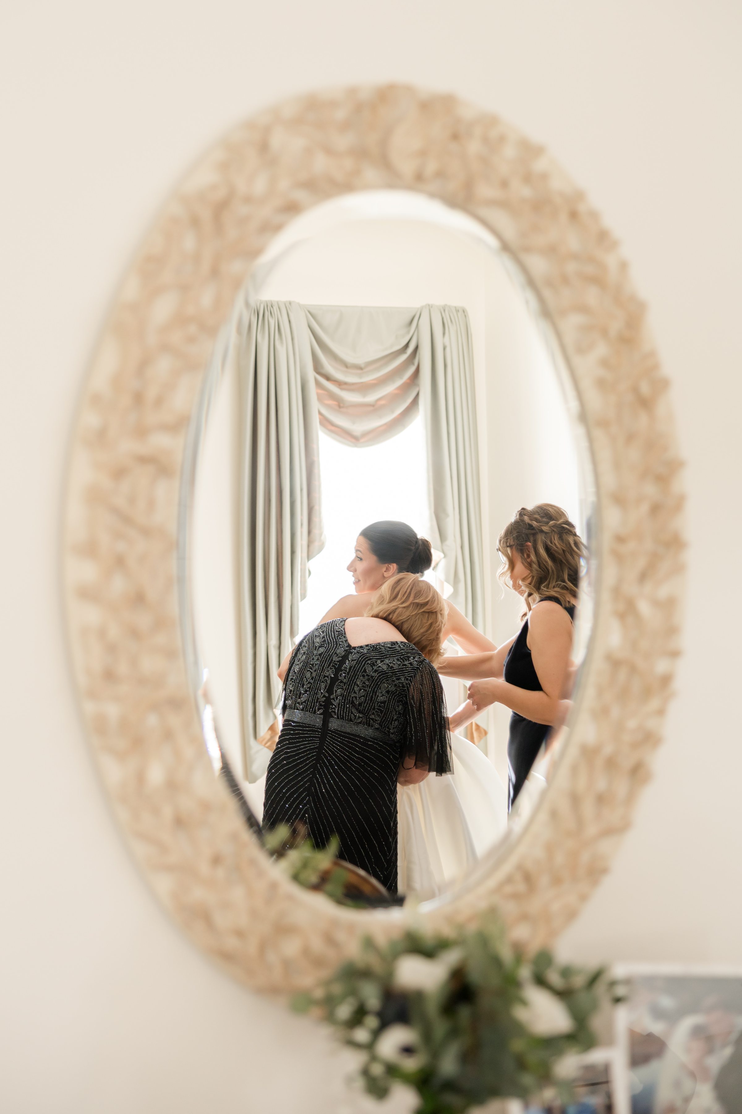 Bride reflected in a mirror as her loved ones help her into her wedding dress, sharing a quiet and heartfelt moment together.