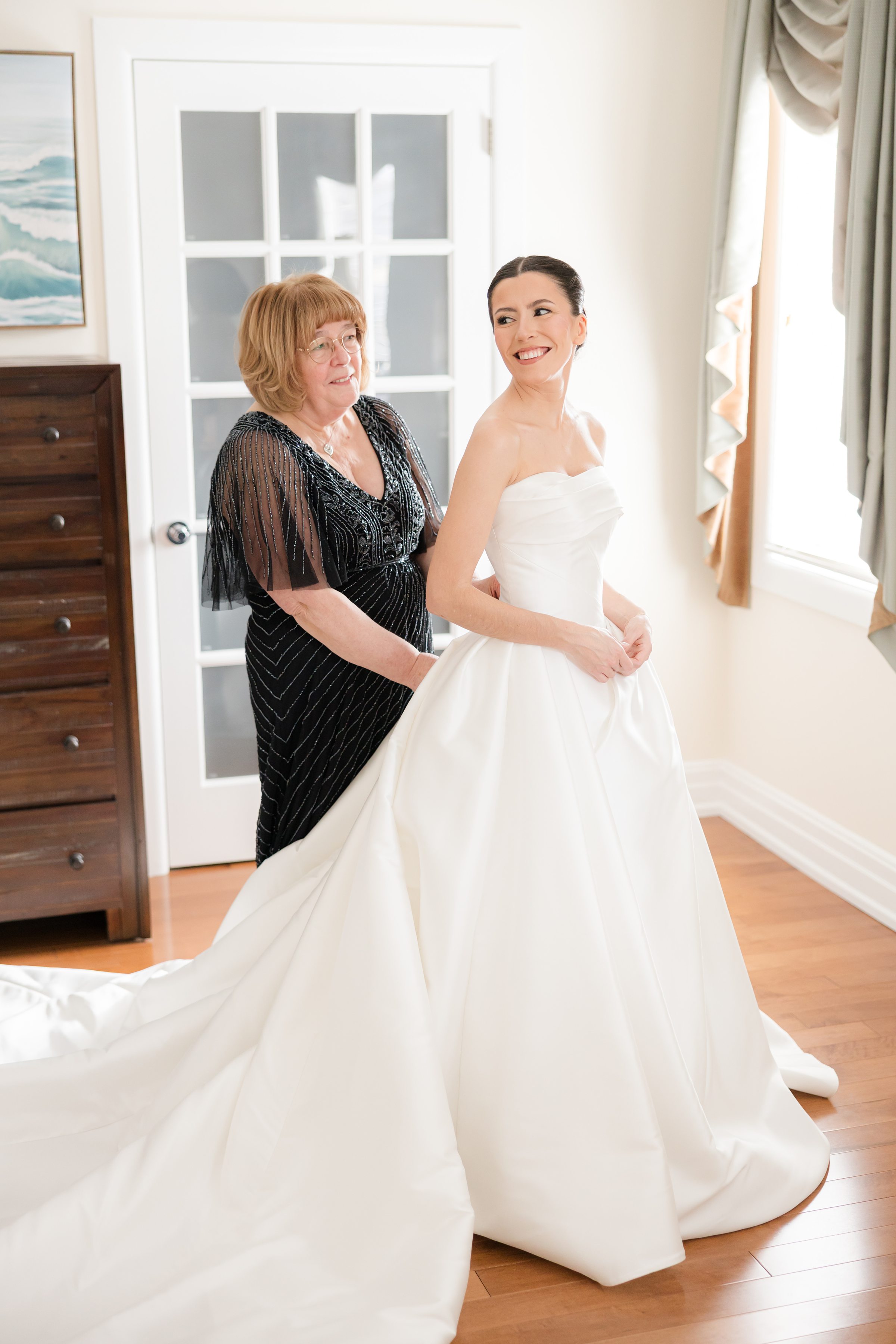 Bride smiling as her mother lovingly fastens the back of her wedding dress, sharing a joyful moment together.