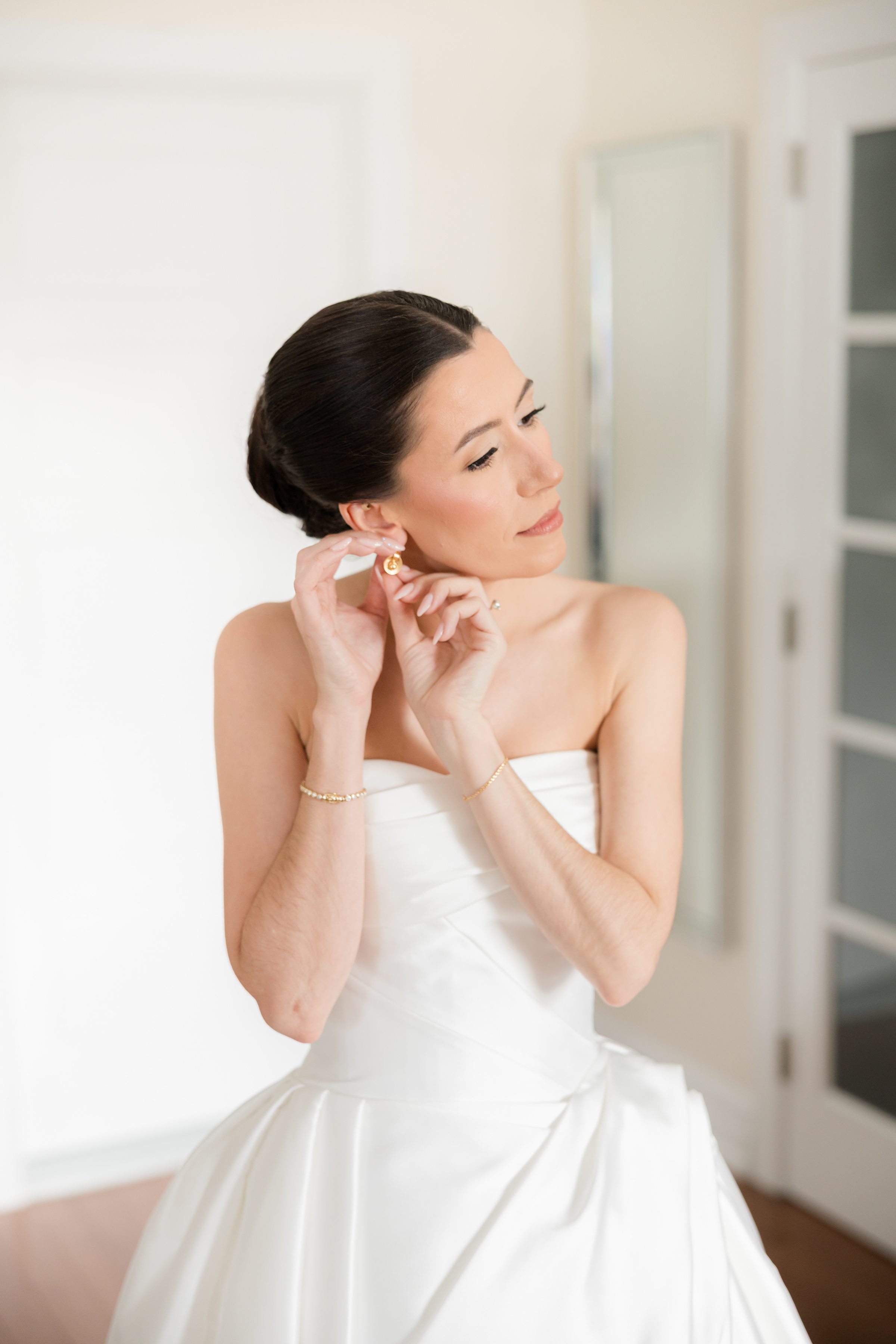 Bride in a strapless white gown adjusts her gold earring, wearing delicate jewelry with her hair in a sleek bun in soft natural light.