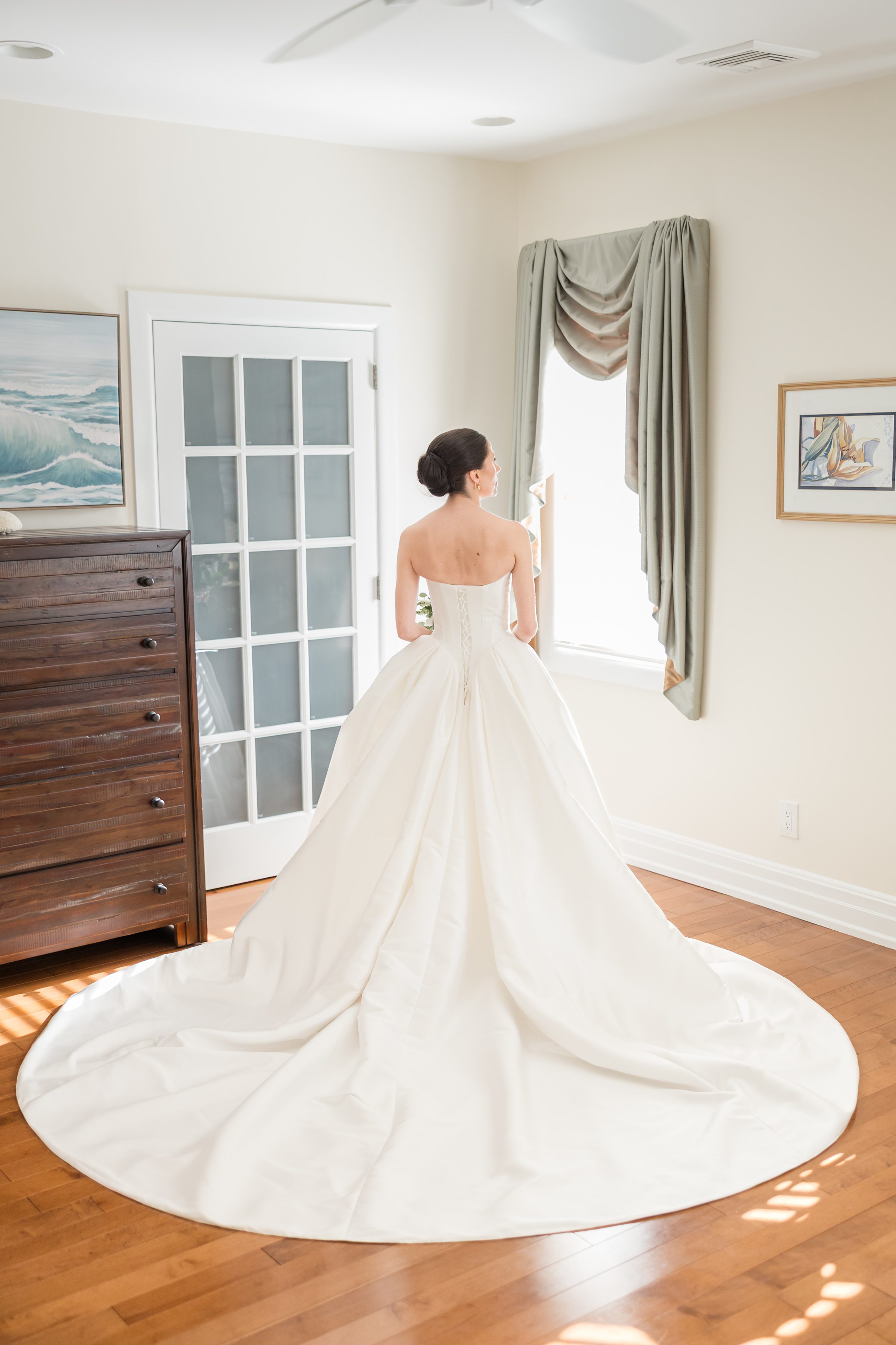 Bride in a full, flowing white gown stands facing a window in a softly lit room with wooden floors, a dresser, and framed artwork.