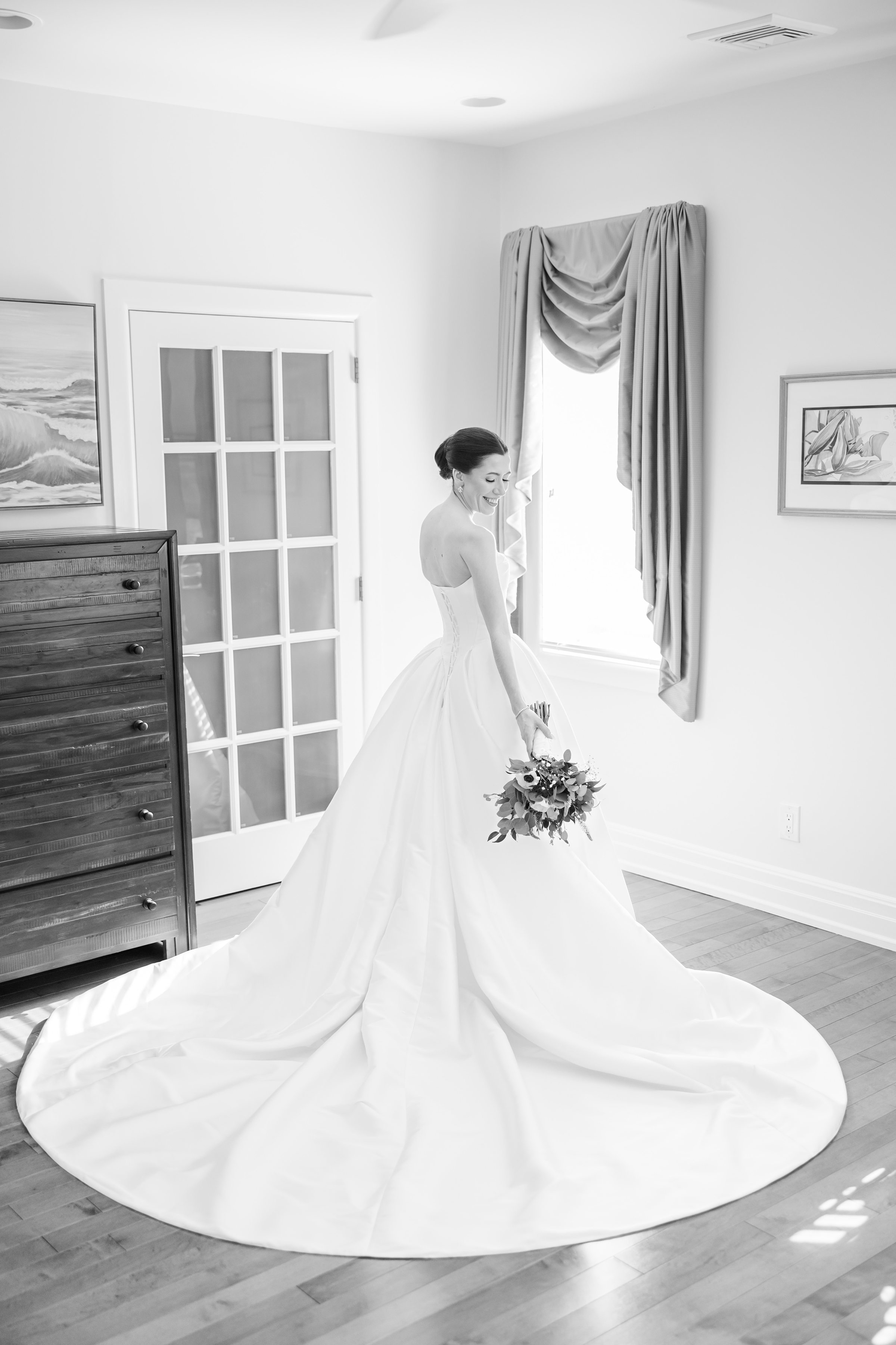Bride in a strapless ball gown holds a bouquet and looks down with a soft smile while standing by a window in a bright room