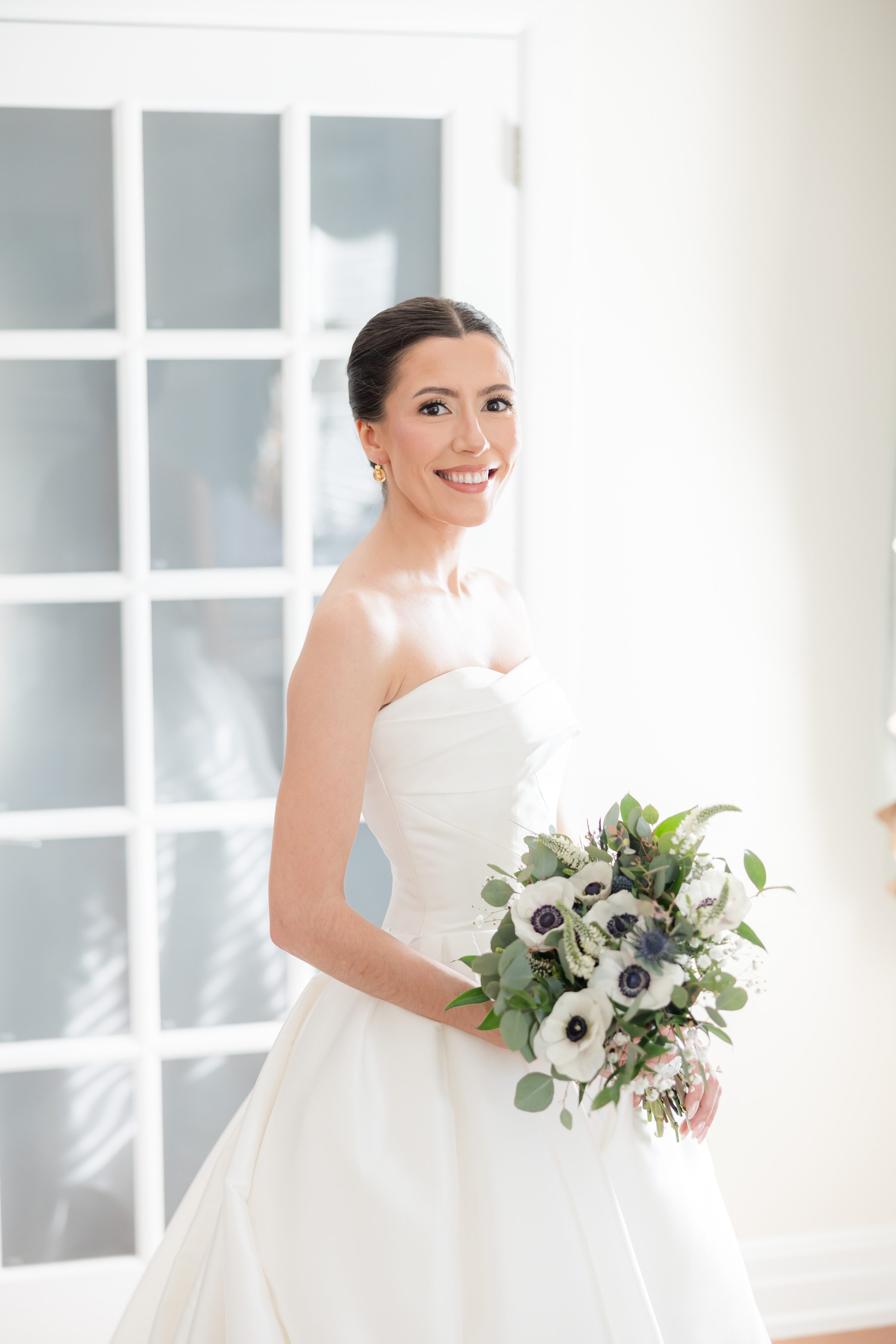 Bride in a strapless white gown smiles while holding a bouquet of greenery and flowers, standing in soft natural light near a glass aneled door.
