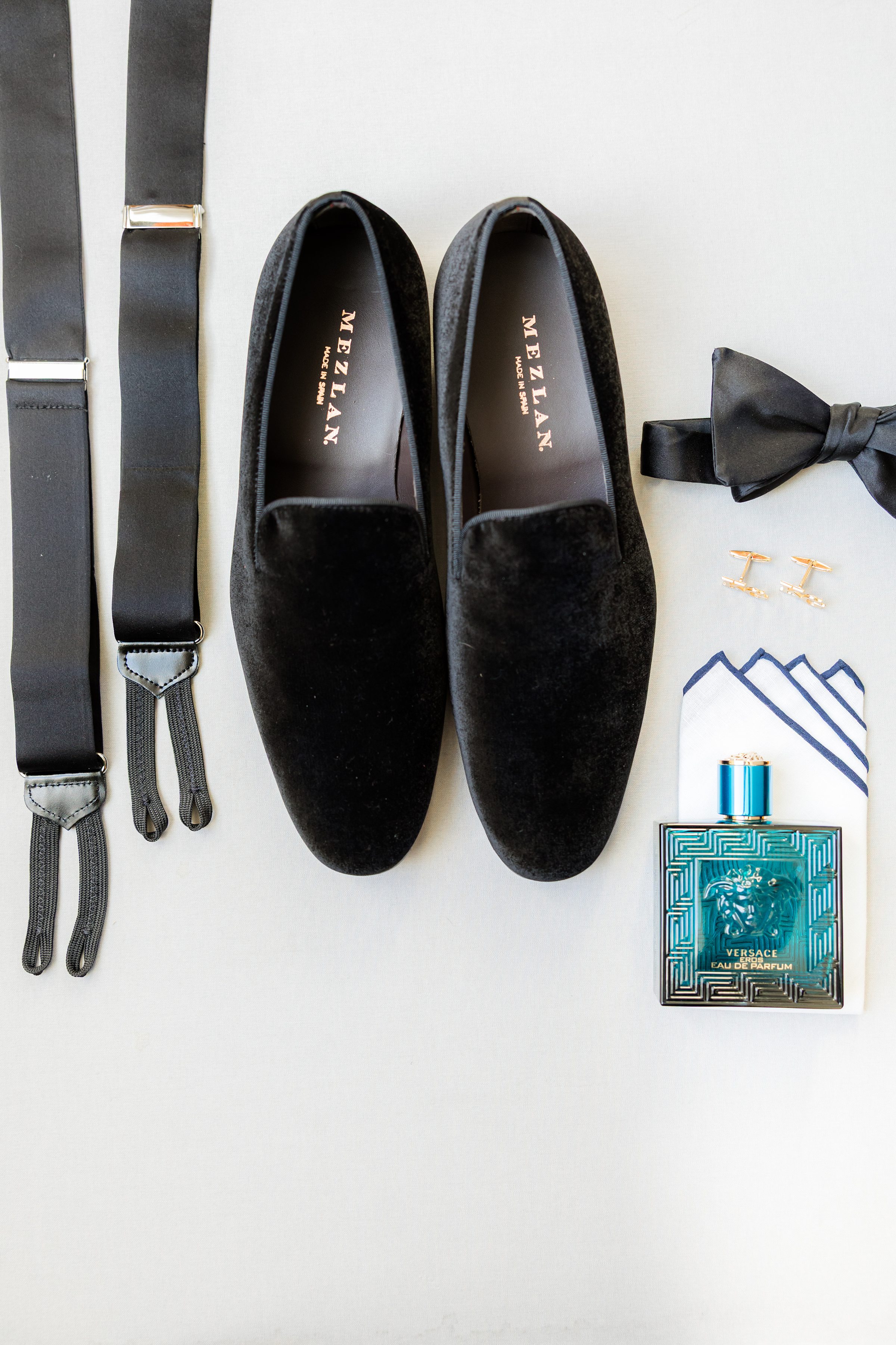 Groom black velvet loafers, suspenders, a bow tie, cufflinks, a pocket square, and a teal cologne bottle arranged neatly on a light surface.