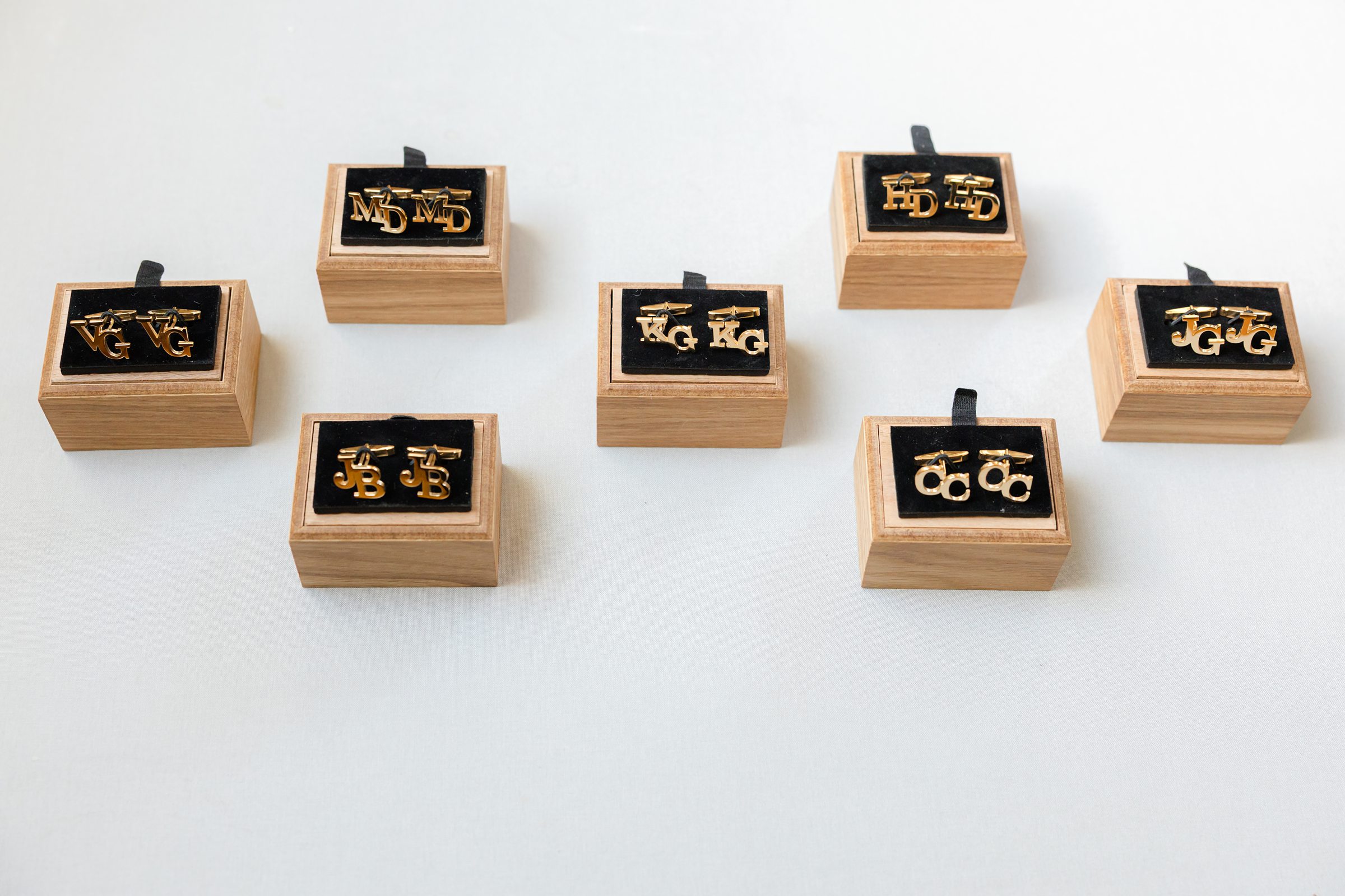 Six pairs of gold cufflinks displayed in small wooden boxes lined with black fabric on a light surface.