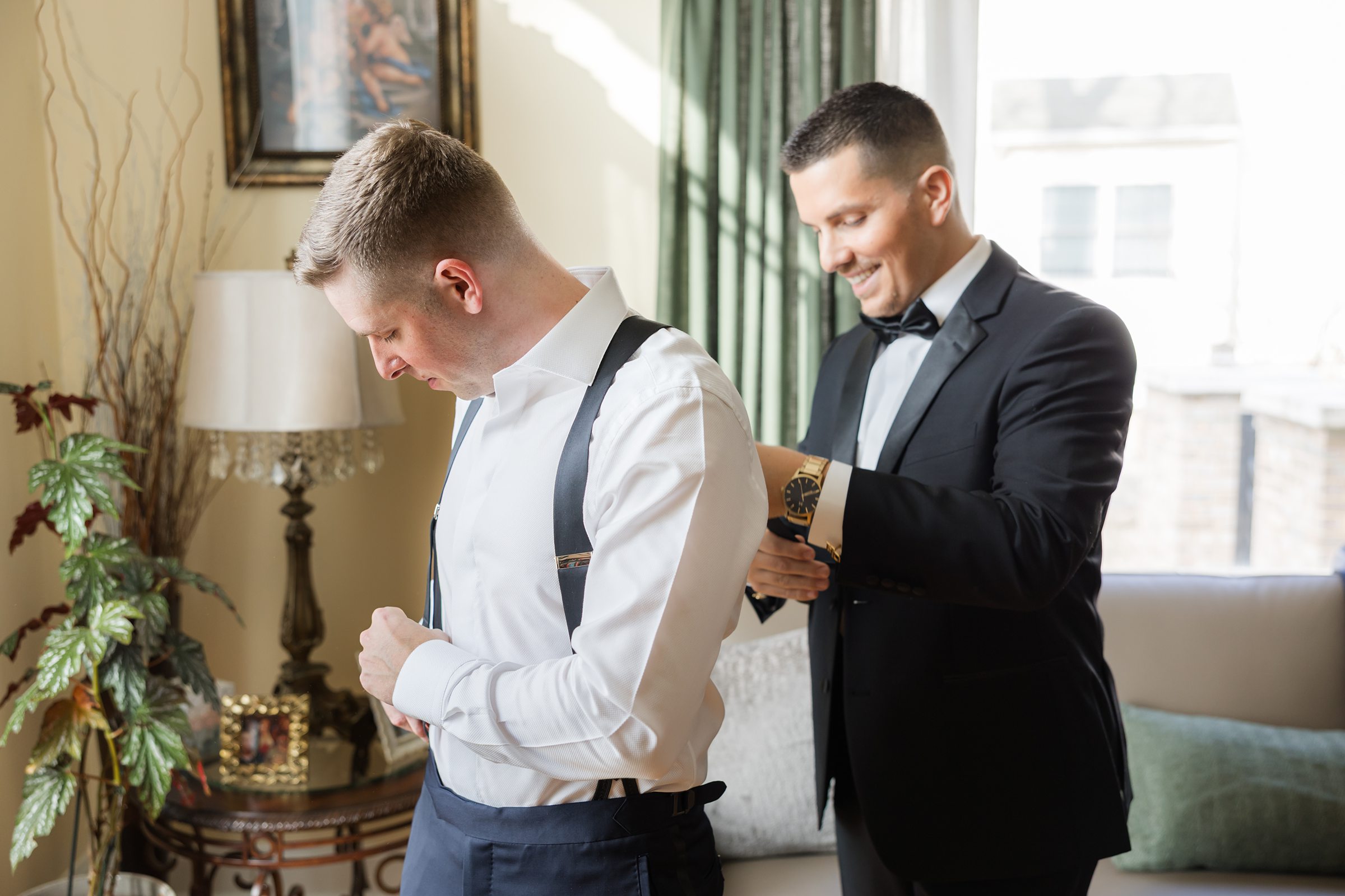 Groom adjusts his suspenders while a groomsman in a tuxedo helps fasten his cuff, standing in a warmly lit room with classic decor.