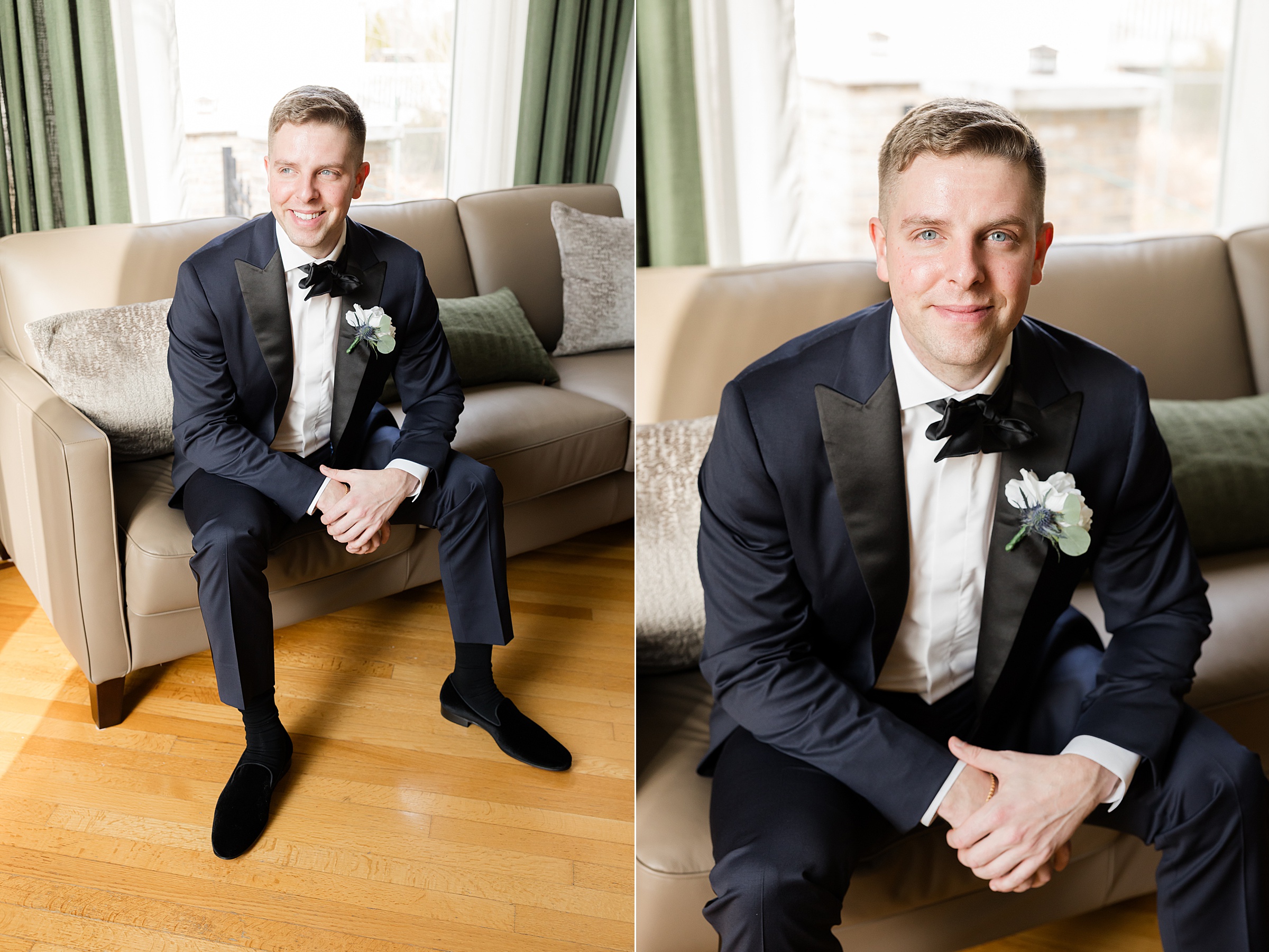 Groom in a black tuxedo sitting on a beige couch by a window, smiling warmly with hands clasped, natural light highlighting his face and boutonniere.