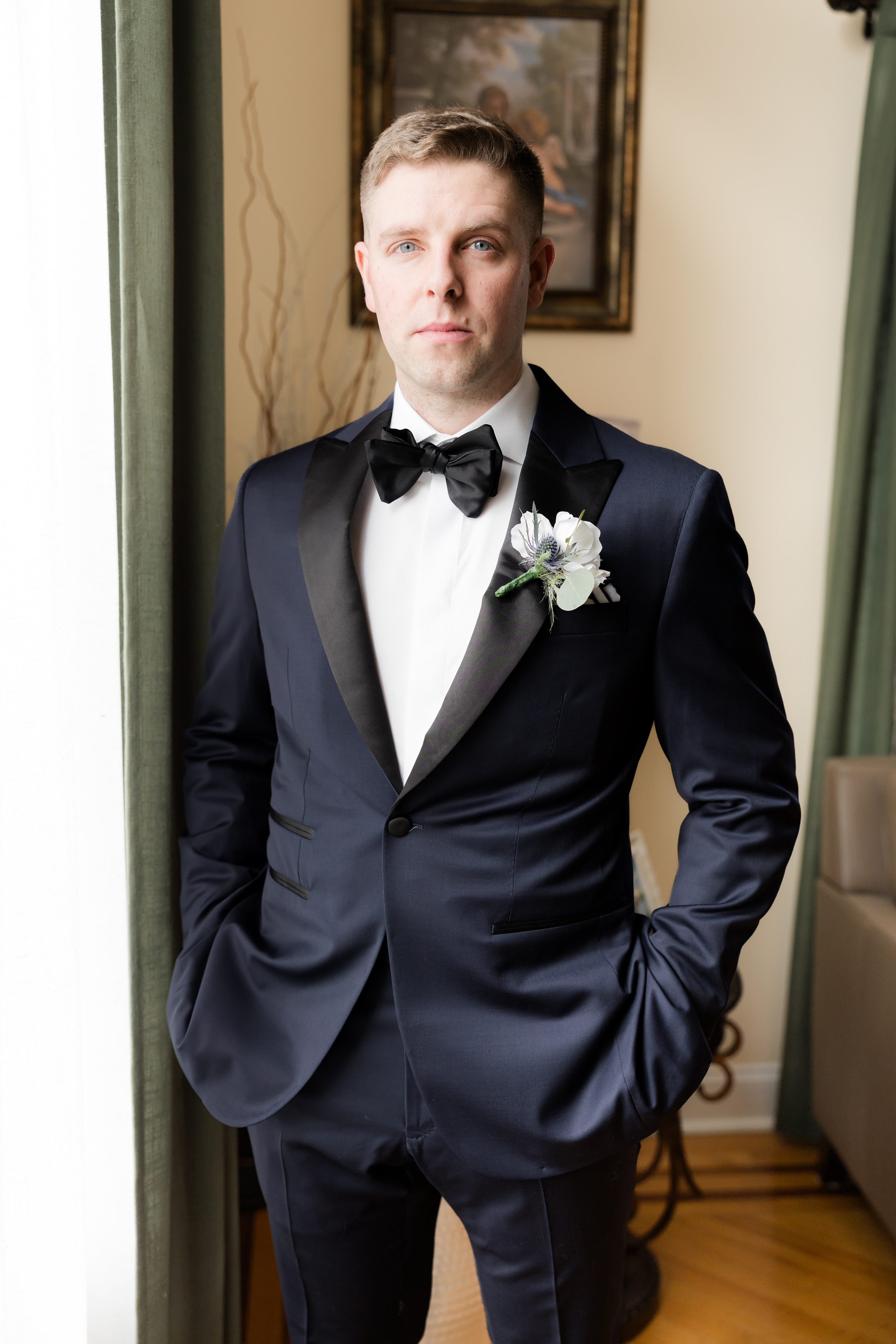 Groom in a black tuxedo with a white boutonniere stands indoors by a window, one hand in his pocket, looking confidently