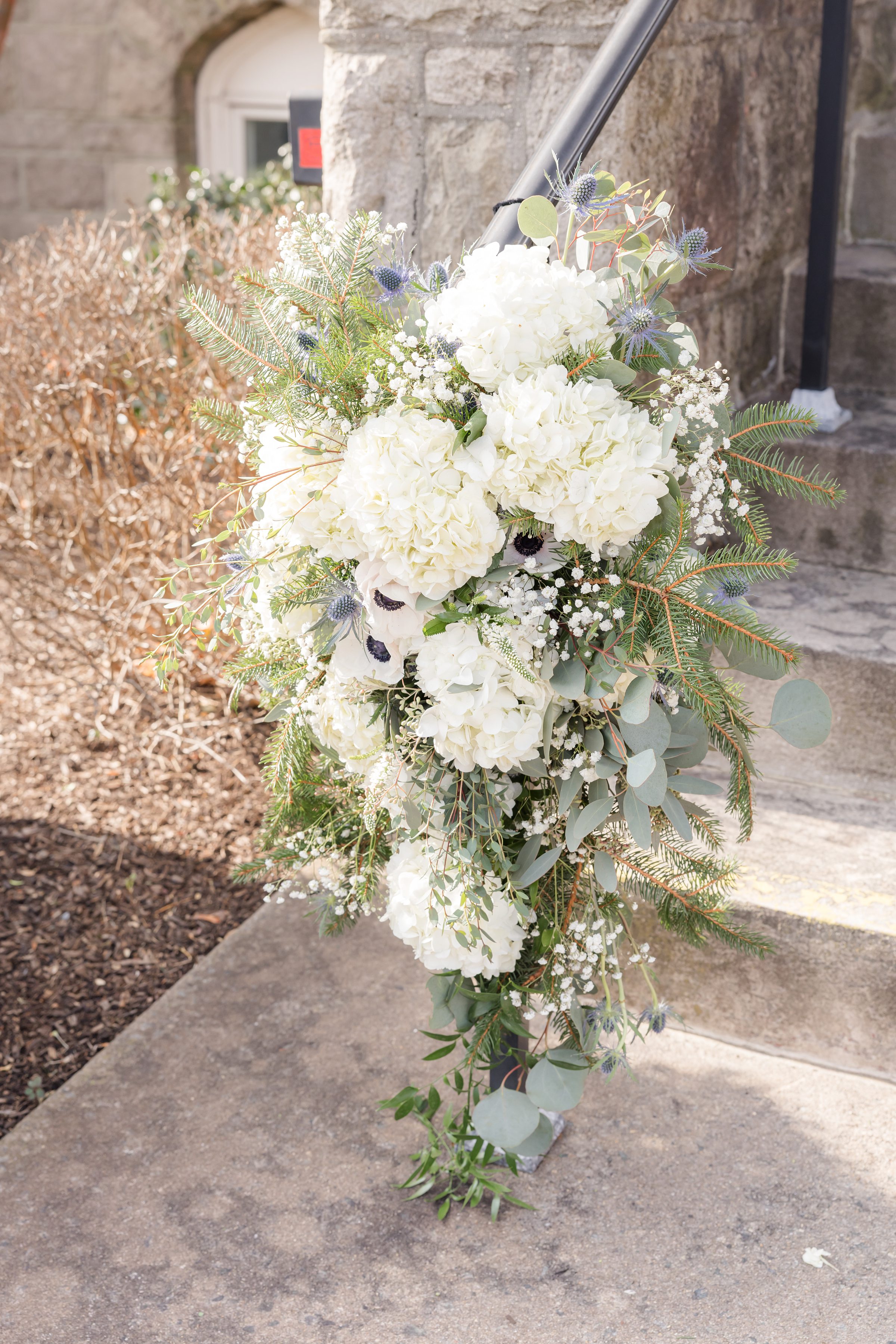 White floral arrangement with hydrangeas, greenery, and small accent flowers decorating a staircase railing outside a stone building.