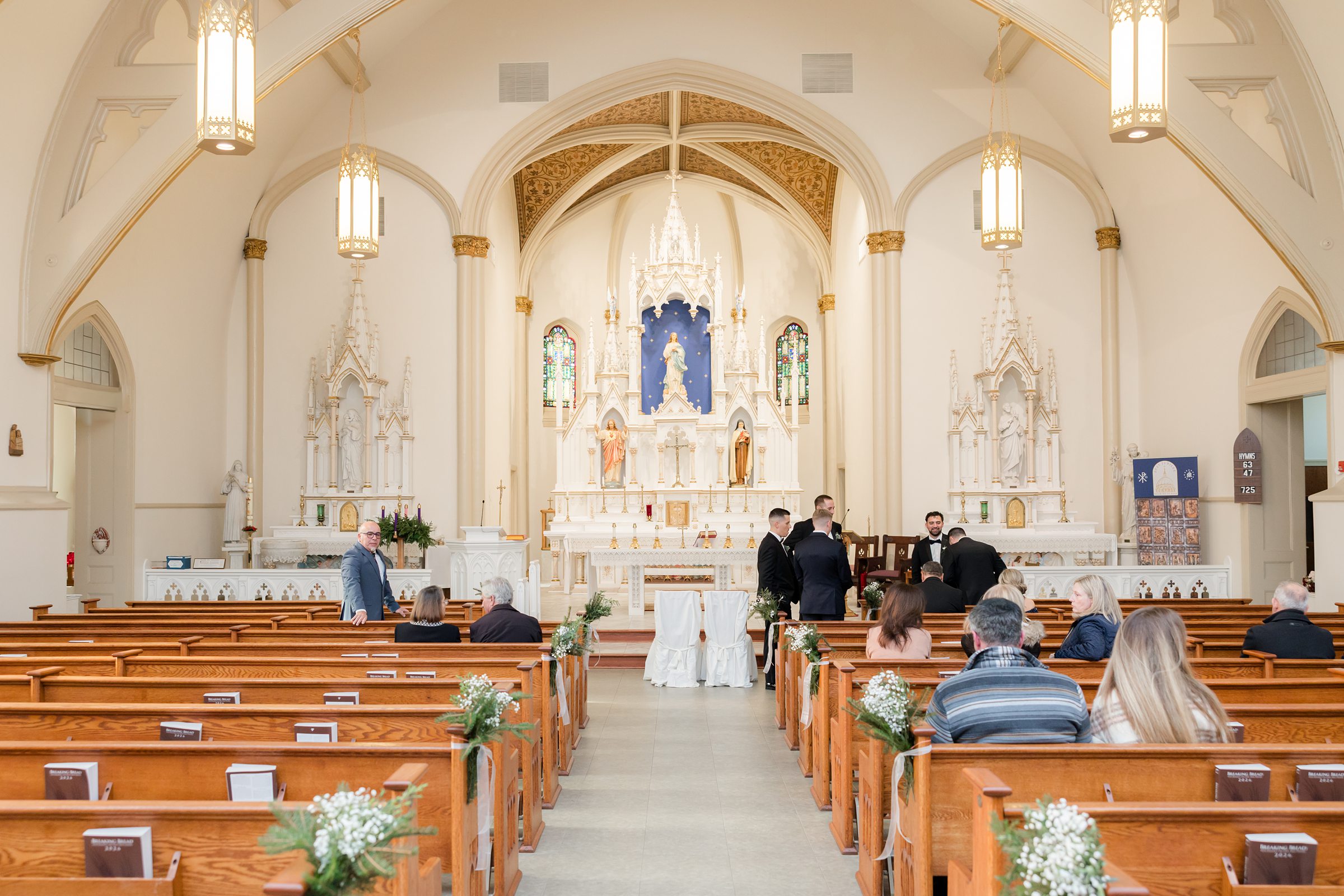 Interior of a bright church with wooden pews decorated with greenery, guests seated, and people gathered near the altar beneath ornate arches and chandeliers.