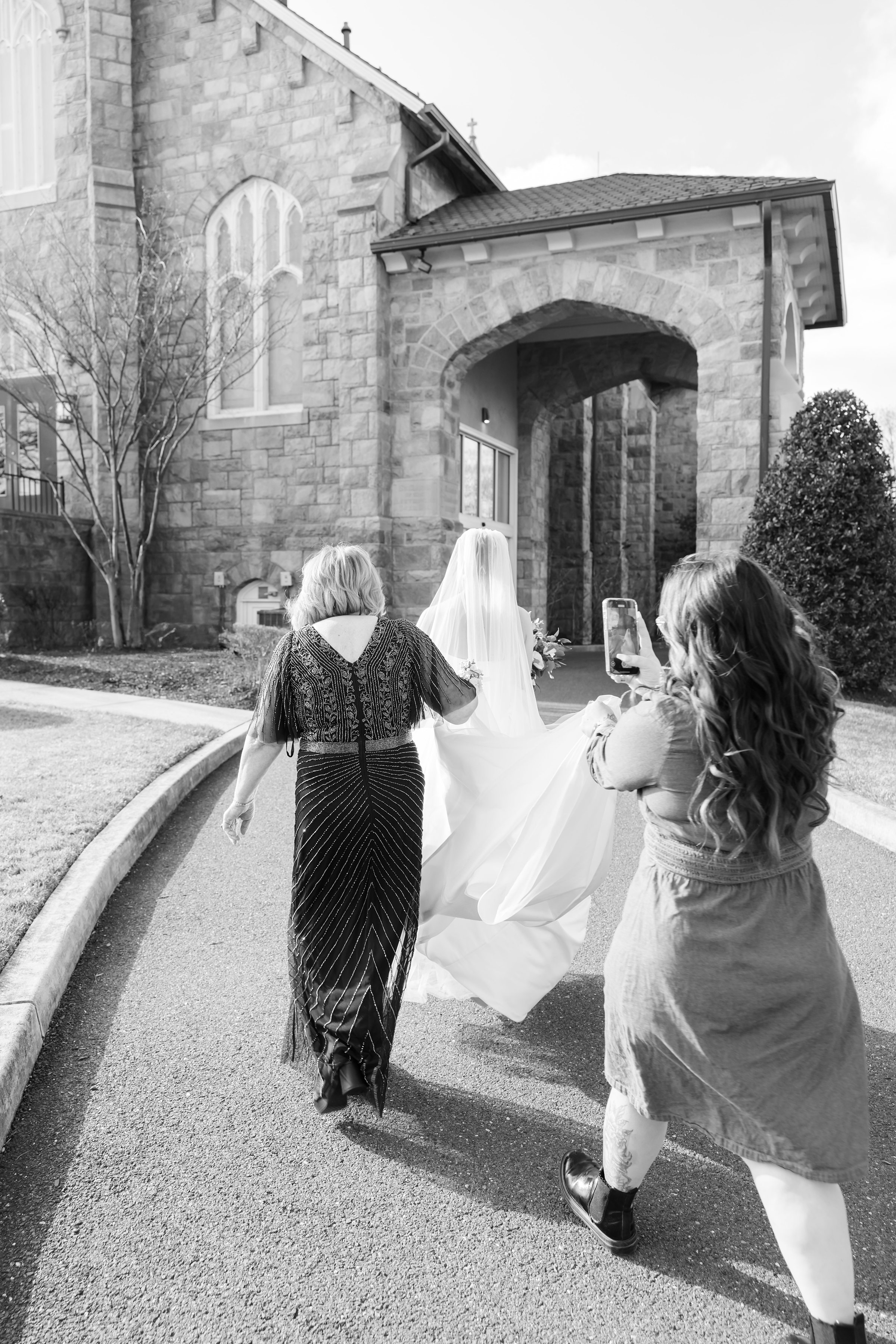 Bride walking outside a stone church with two loved ones, her veil and dress flowing as one person holds her gown and another records the moment on a phone.