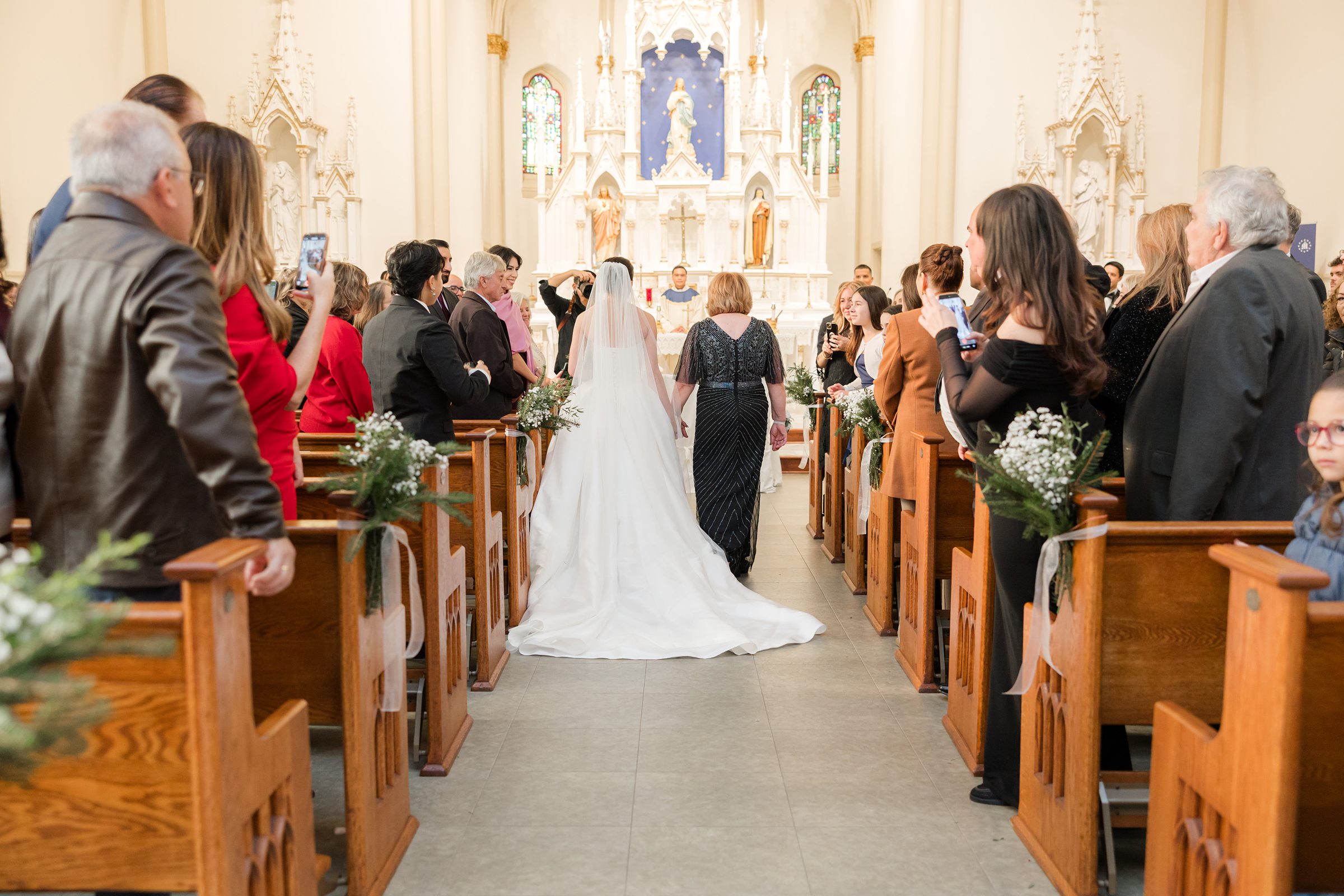 Bride walking down the church aisle with her mother, guests standing on both sides of decorated pews as they approach the altar.