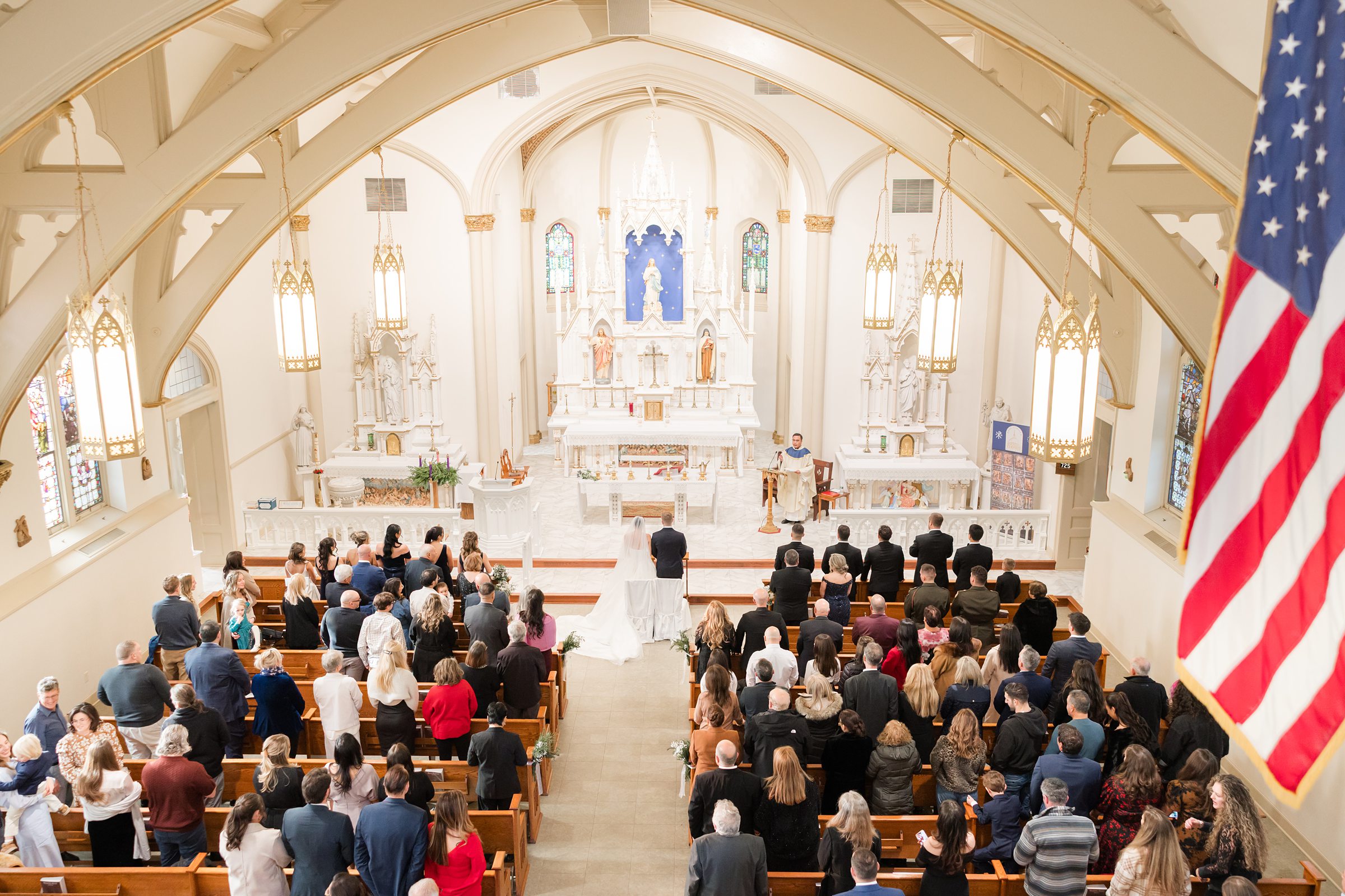 Wedding ceremony in a church, with the bride and groom standing at the altar as guests fill the pews beneath vaulted ceilings and hanging lights.