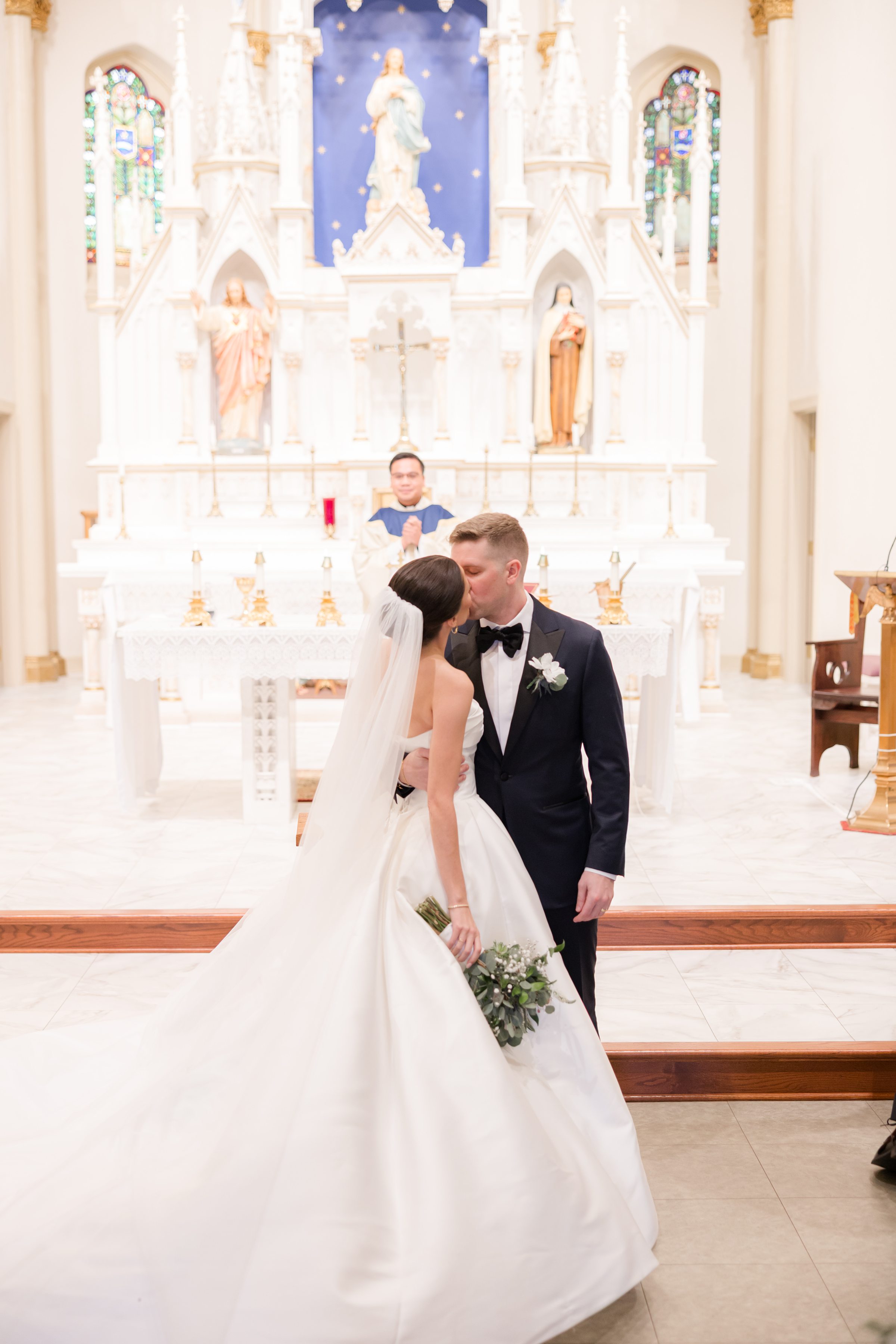 Bride and groom share a kiss at the altar during their wedding ceremony, with the officiant and ornate church backdrop behind them.