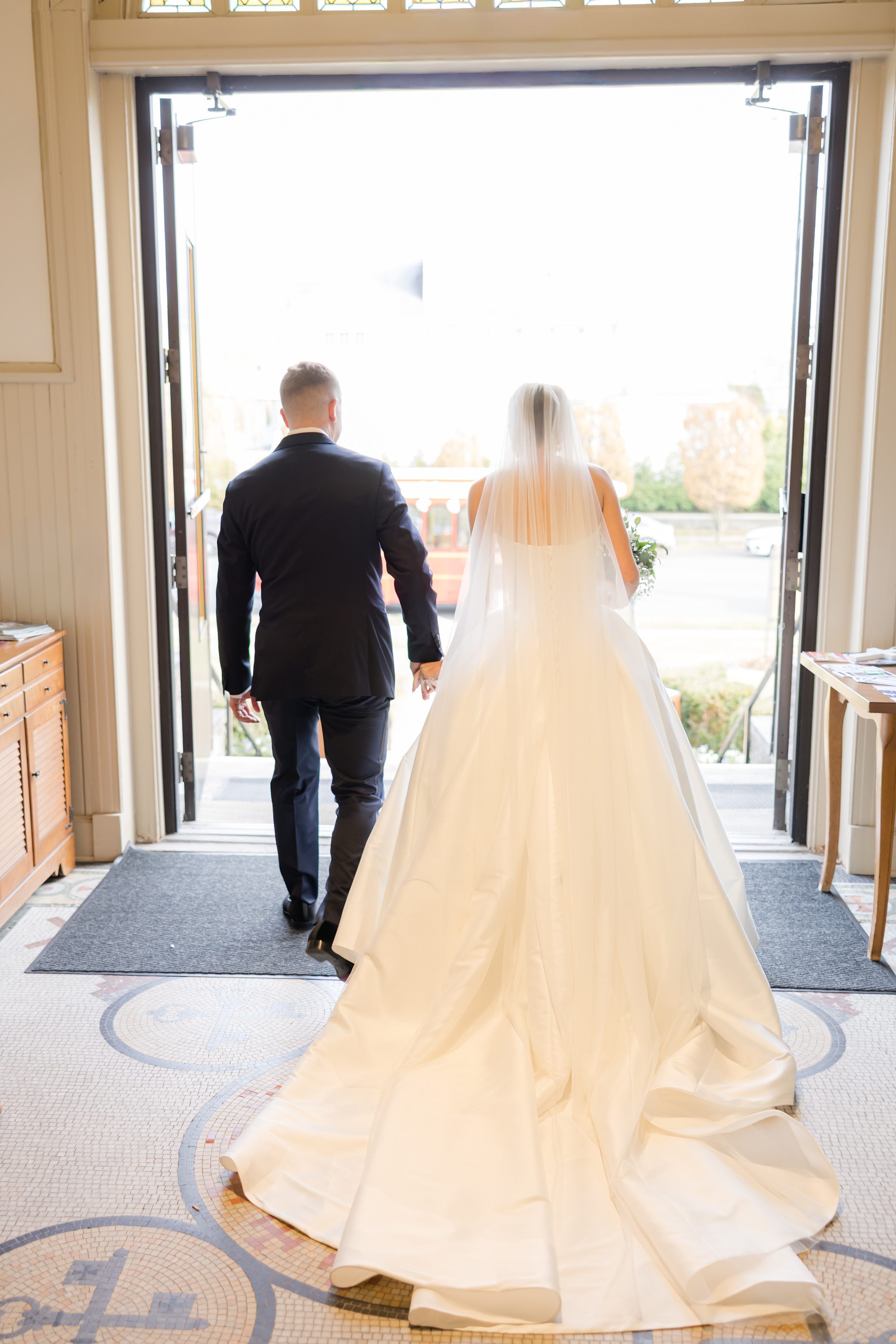 Bride and groom holding hands from behind, walking out of church doors into bright daylight.