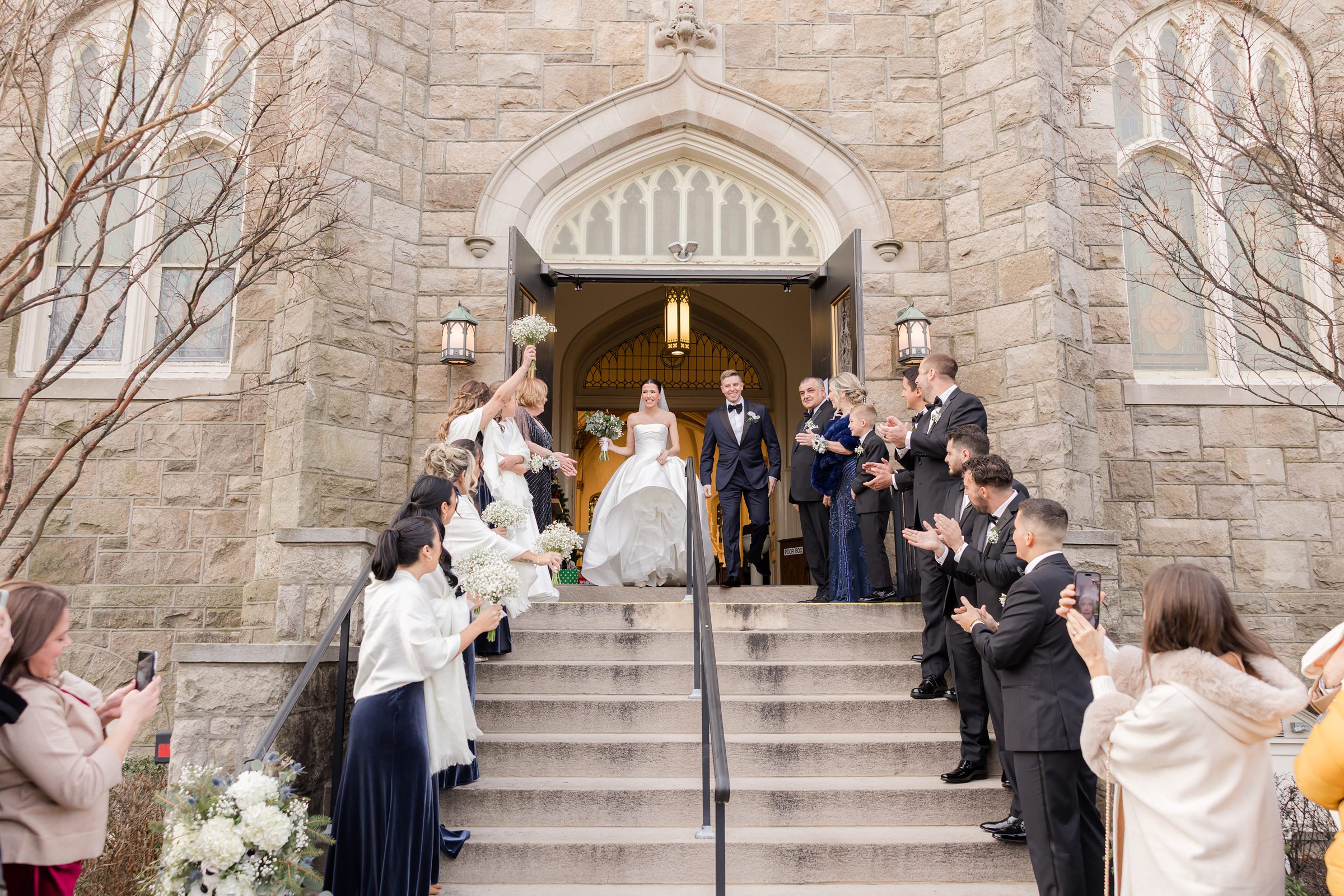 Bride and groom exiting a stone church while guests line the steps celebrating.