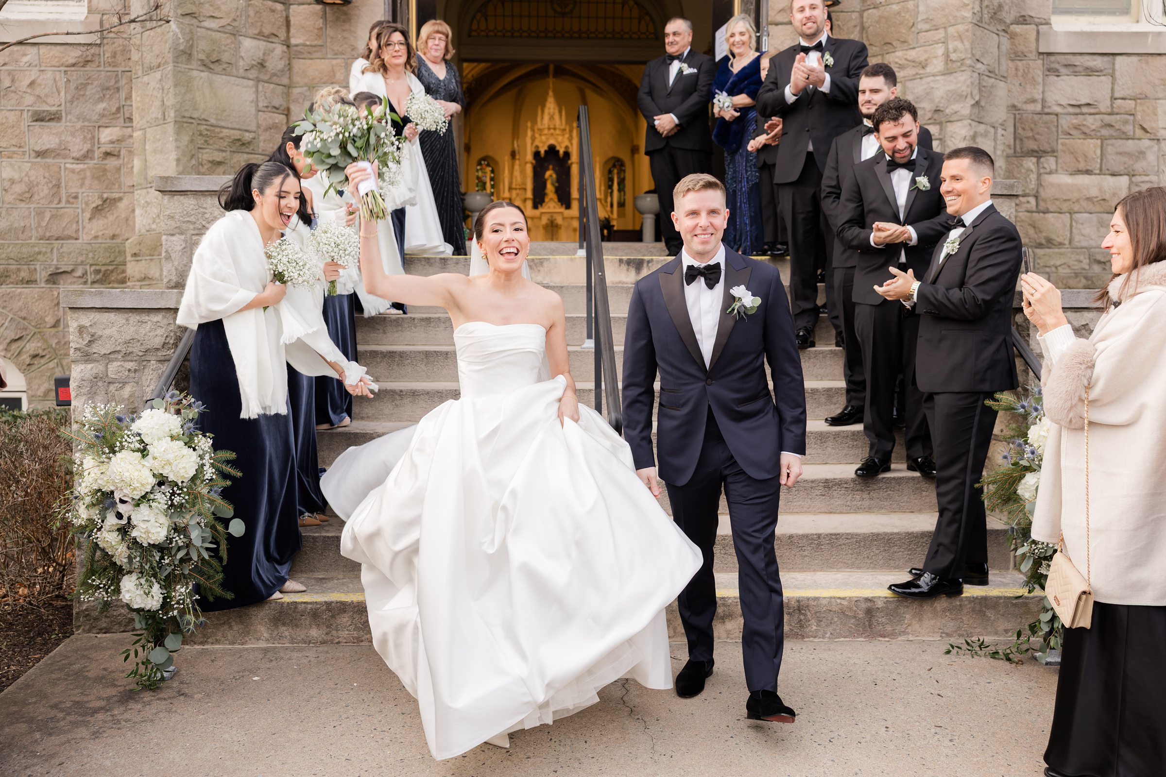 Bride and groom smiling and walking down church steps as wedding party cheers and throws flowers.