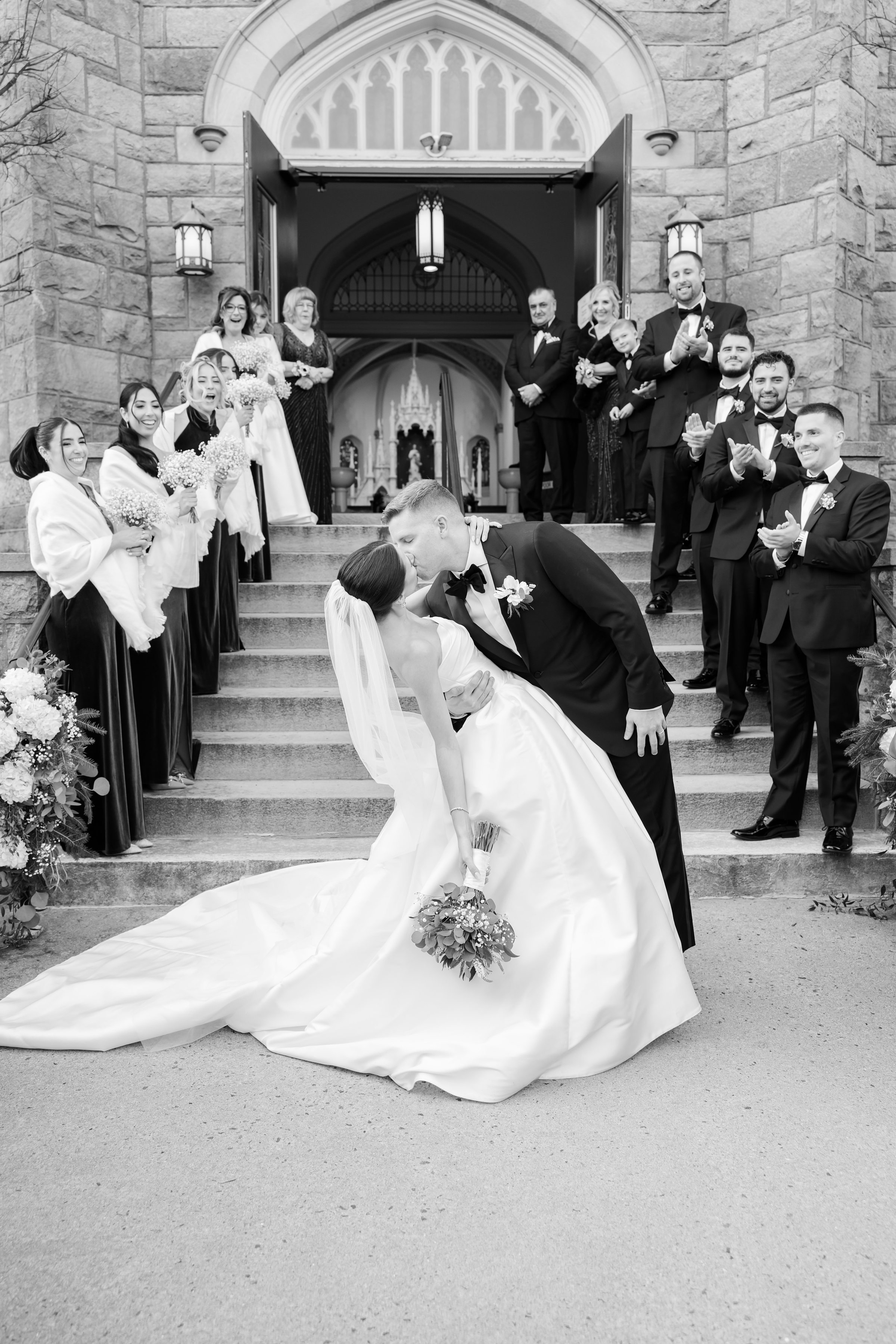 Bride and groom sharing a dip kiss outside a church, surrounded by cheering wedding guests.