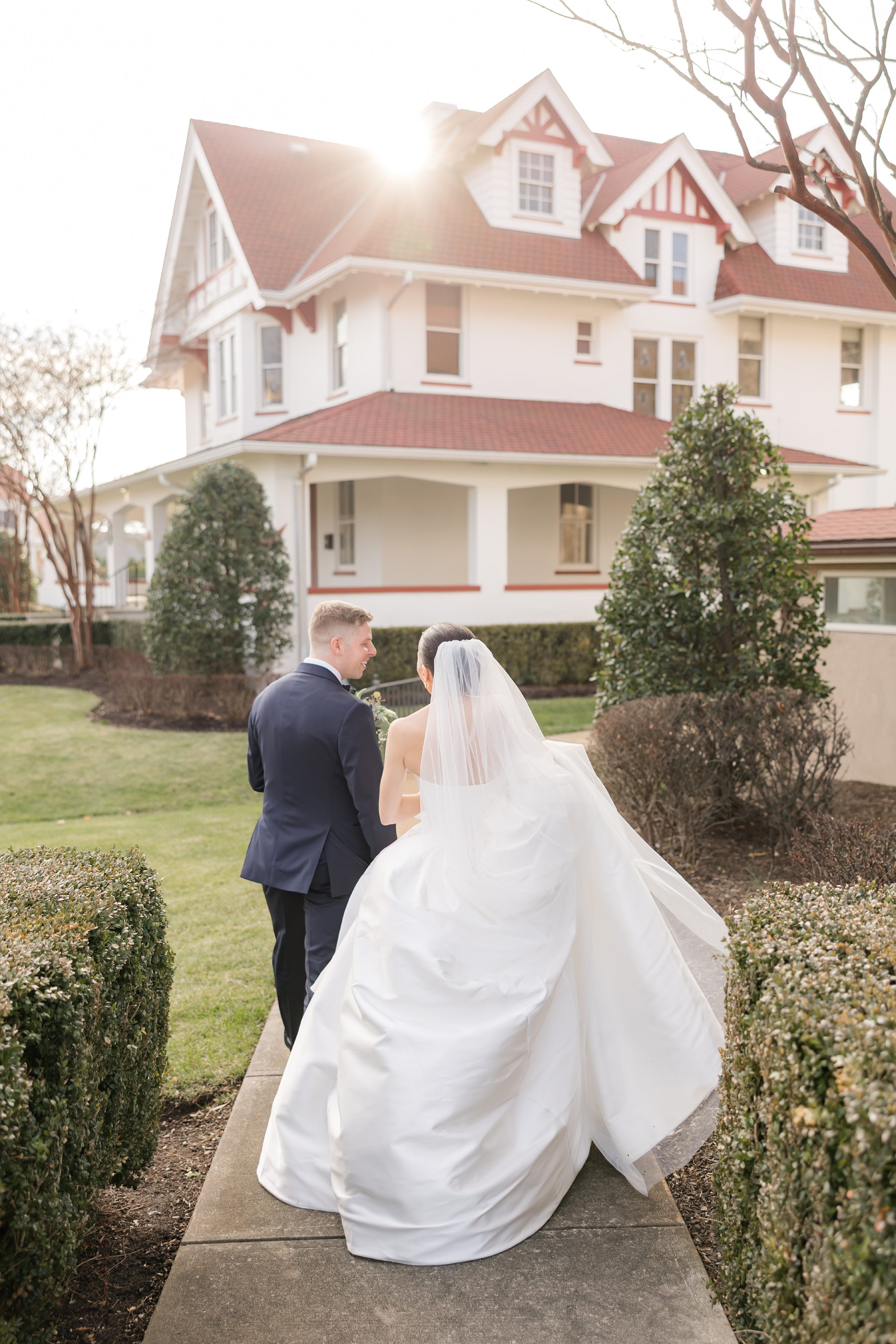 Bride and groom walking away together along a path toward a large house, sunlight glowing behind them.