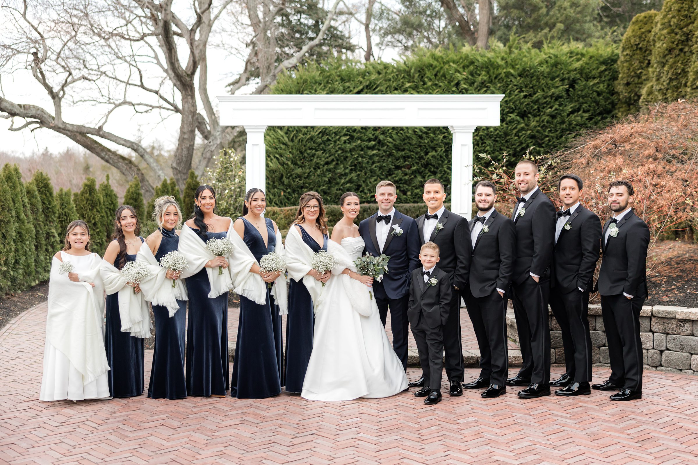 Wedding party portrait with bride and groom centered, surrounded by bridesmaids in navy dresses and groomsmen in black tuxedos, posing together under a white garden arbor on a brick courtyard.