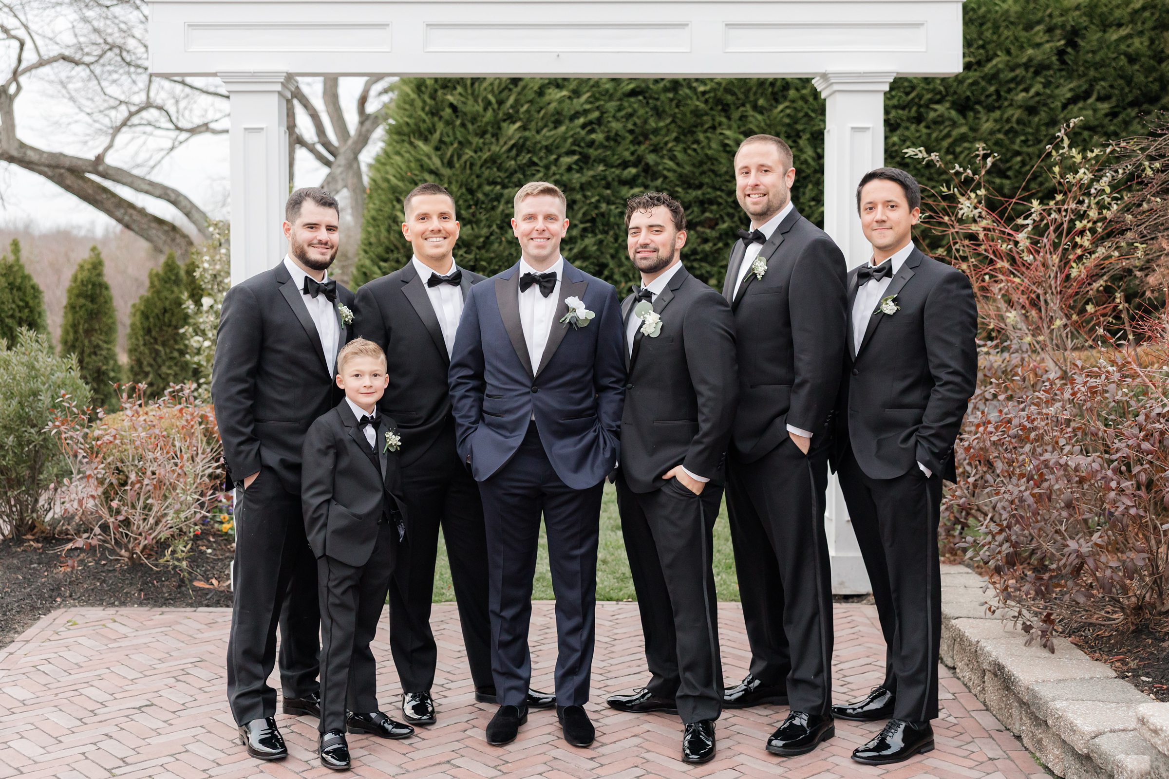 Groom with groomsmen and a young boy, all dressed in black tuxedos, posing outdoors.