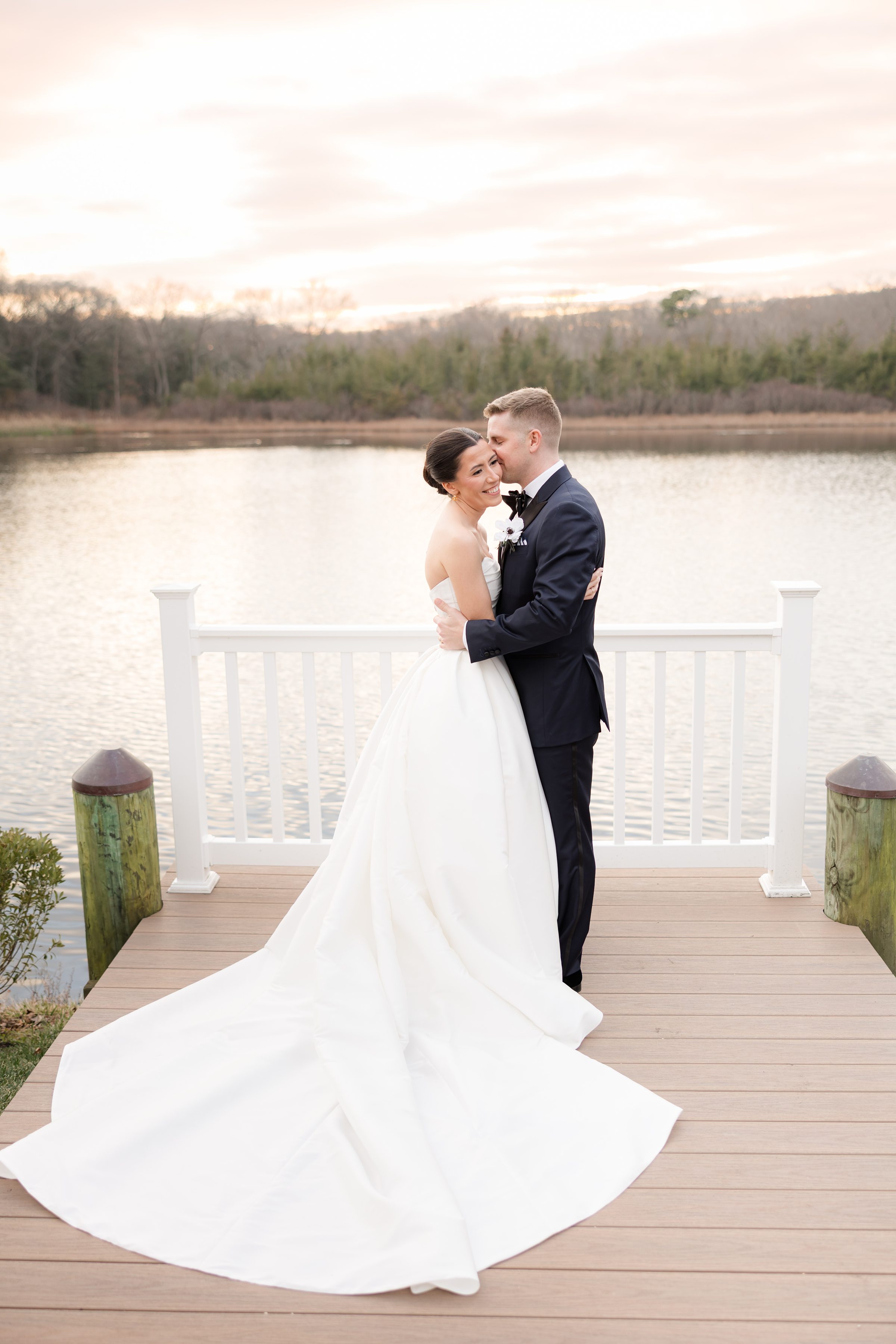 Groom kissing bride’s cheek as she smiles, both standing on a dock at sunset.