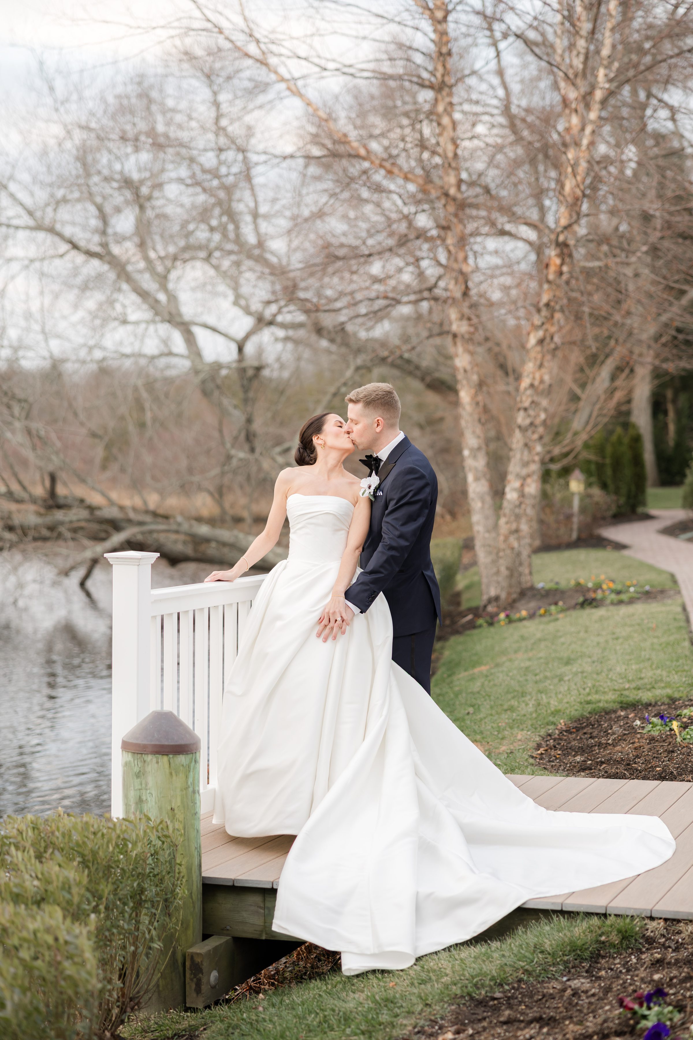 Bride and groom kissing by a lakeside railing with trees in the background.