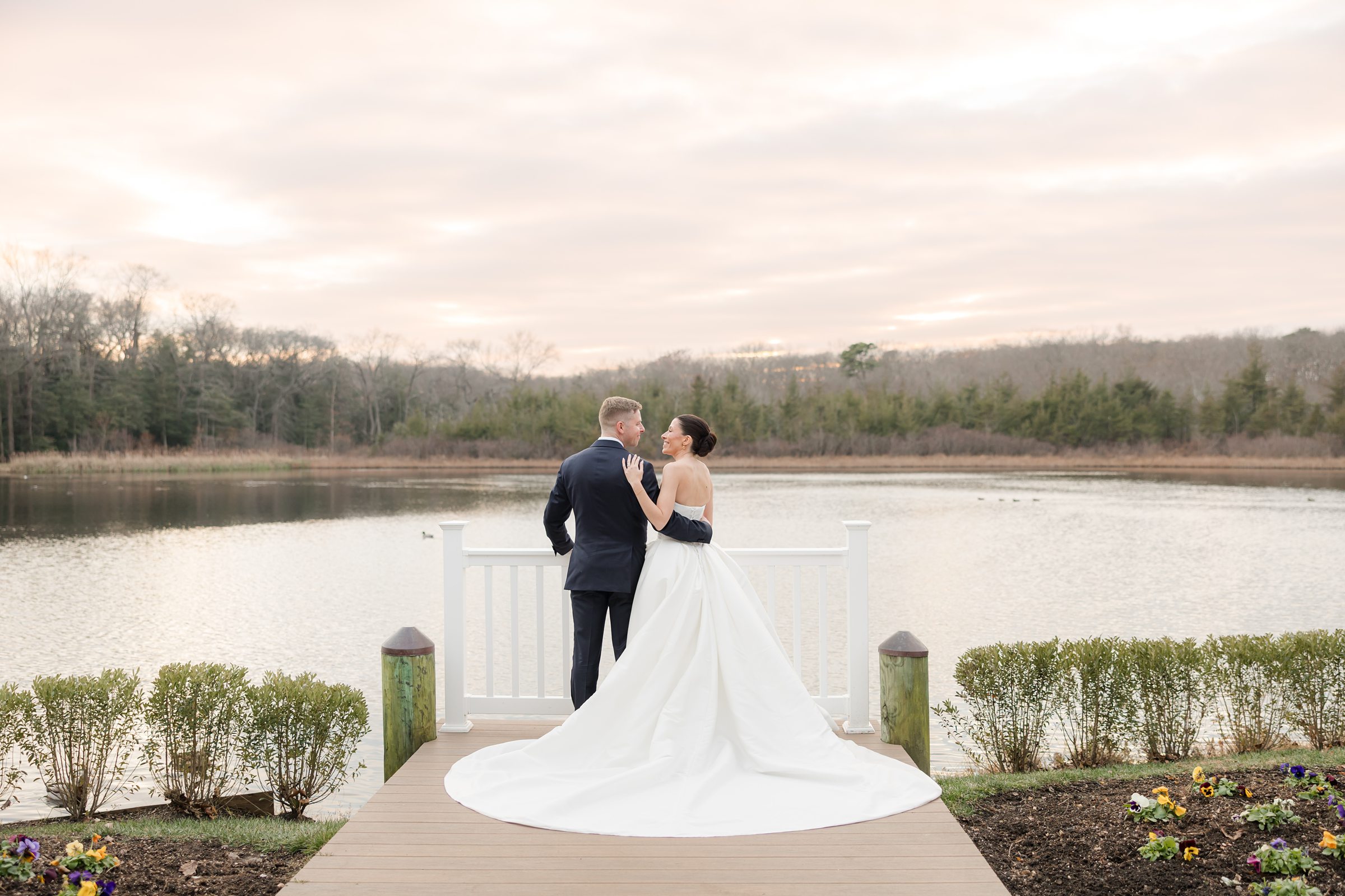 Bride and groom stand close together at the end of a wooden dock, gazing at each other with a calm lake and soft sunset sky behind them, her gown flowing elegantly across the boards.