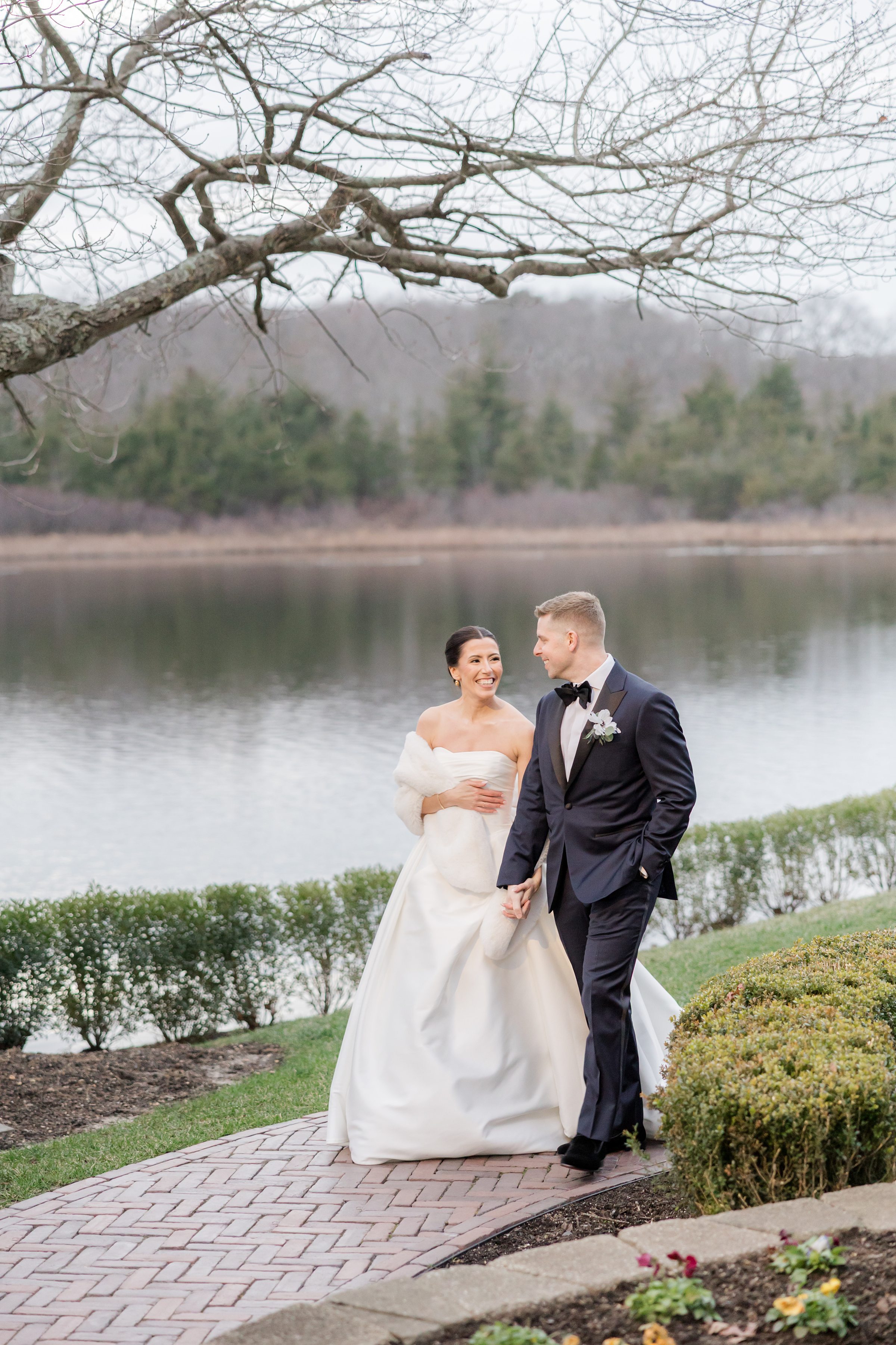 Bride and groom holding hands and walking along a curved brick path beside a calm lake.