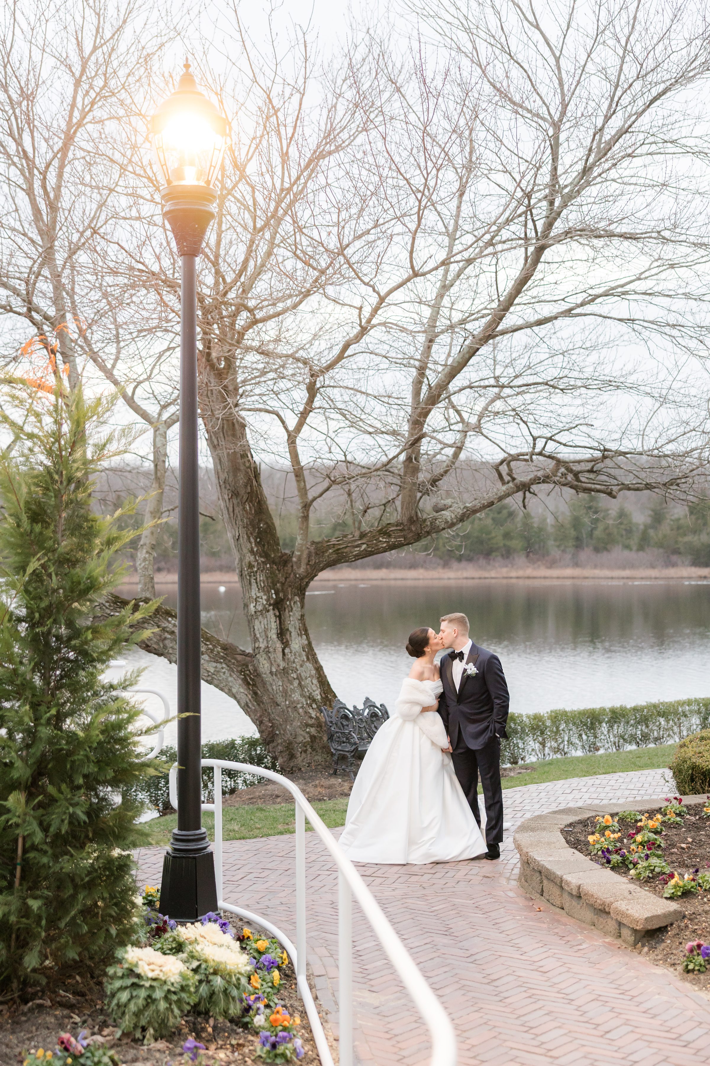 Bride and groom share a tender kiss by the lakeside beneath a glowing lamppost, framed by bare trees and soft, peaceful light.