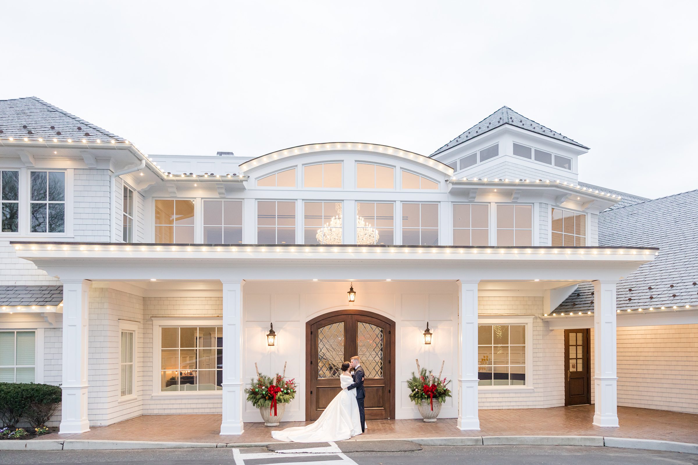 Bride and groom share a kiss under the covered entrance of a large, elegant white house with lit windows and floral arrangements.