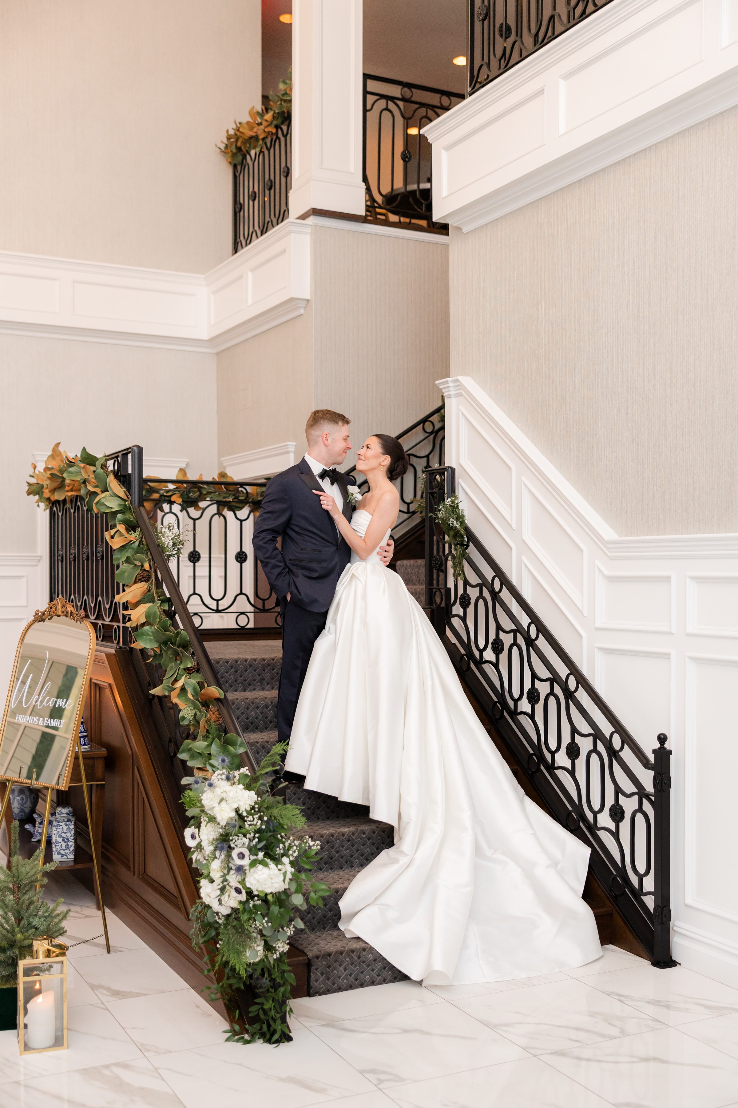 Bride and groom standing on a grand staircase decorated with greenery and white flowers, gazing at each other.