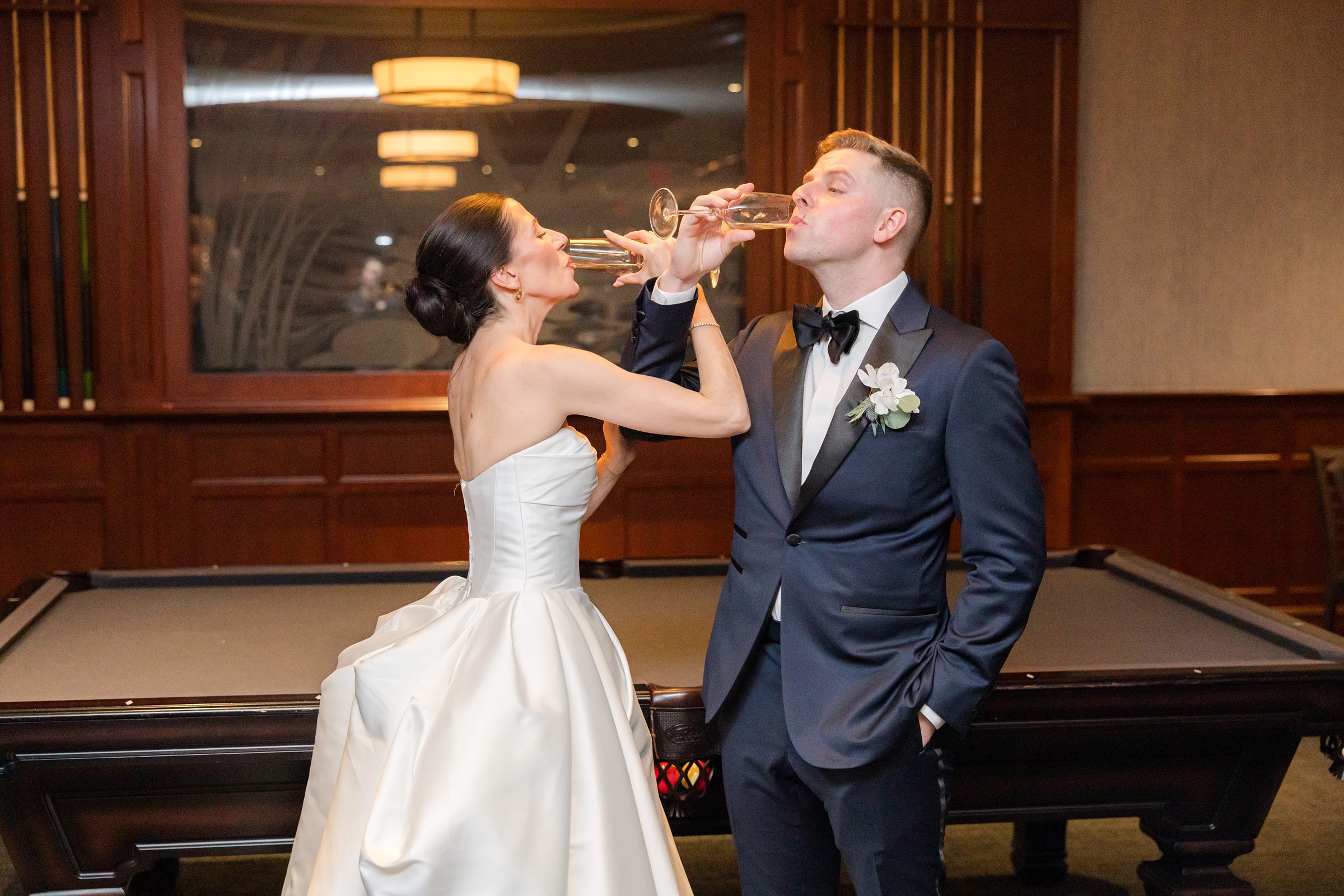 Bride and groom drinking champagne together in a lounge with a pool table behind them.