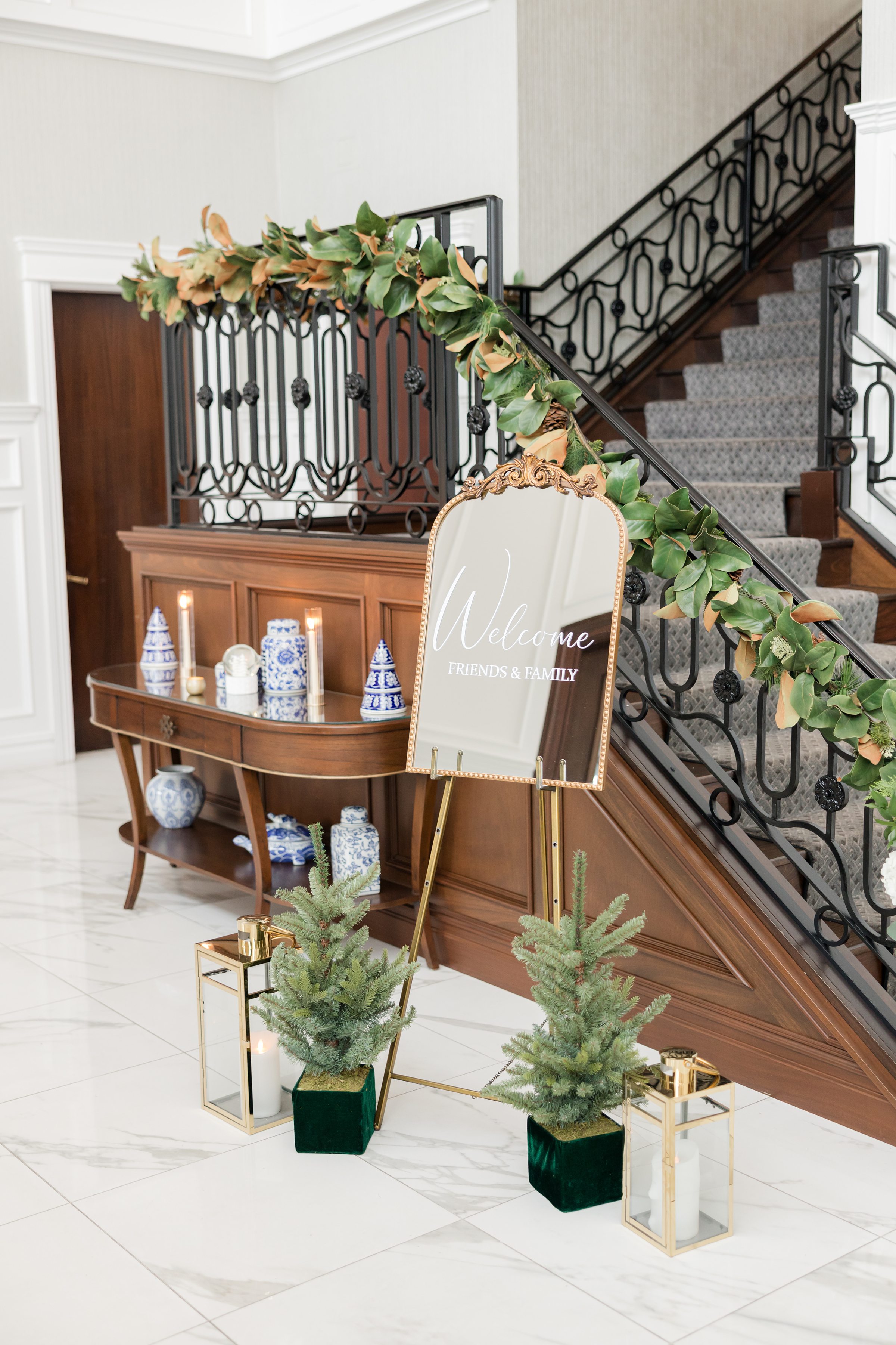 Wedding welcome sign on an easel, surrounded by greenery, candles, and decorative accents beside a staircase with garland.