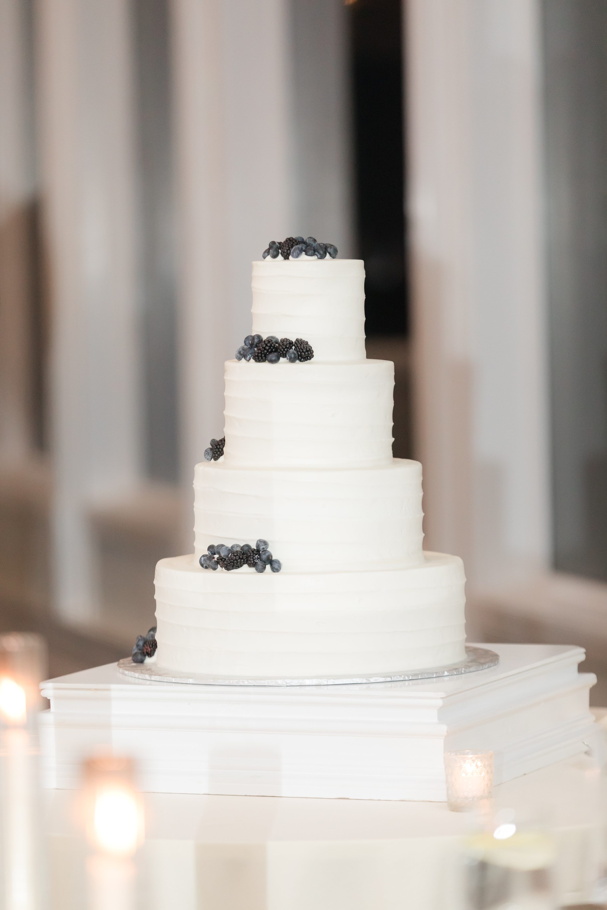 Elegant four-tier white wedding cake decorated with blueberries and blackberries, displayed on a white pedestal.