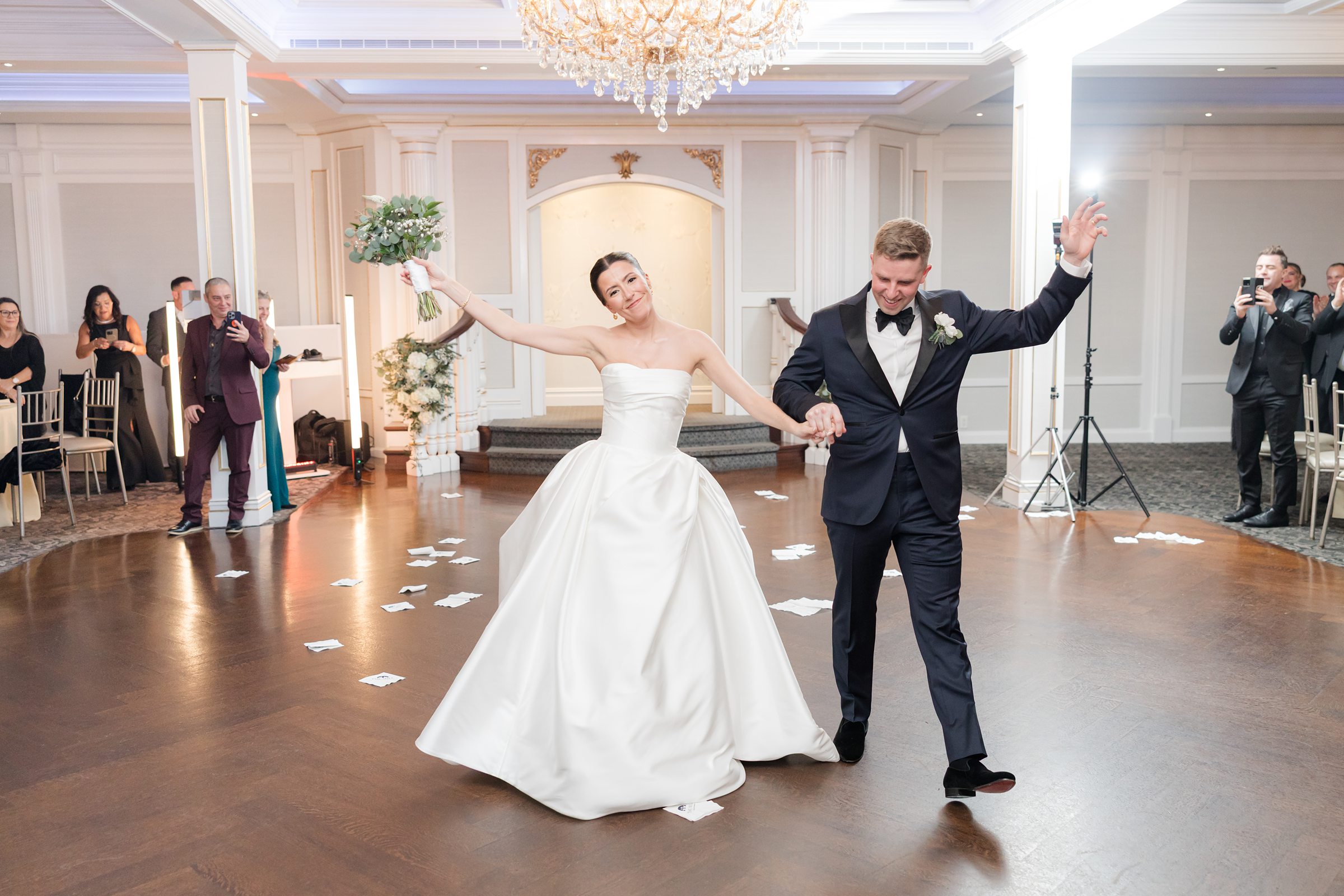 Bride and groom celebrating on the dance floor, holding hands with arms raised, while guests watch in the background.