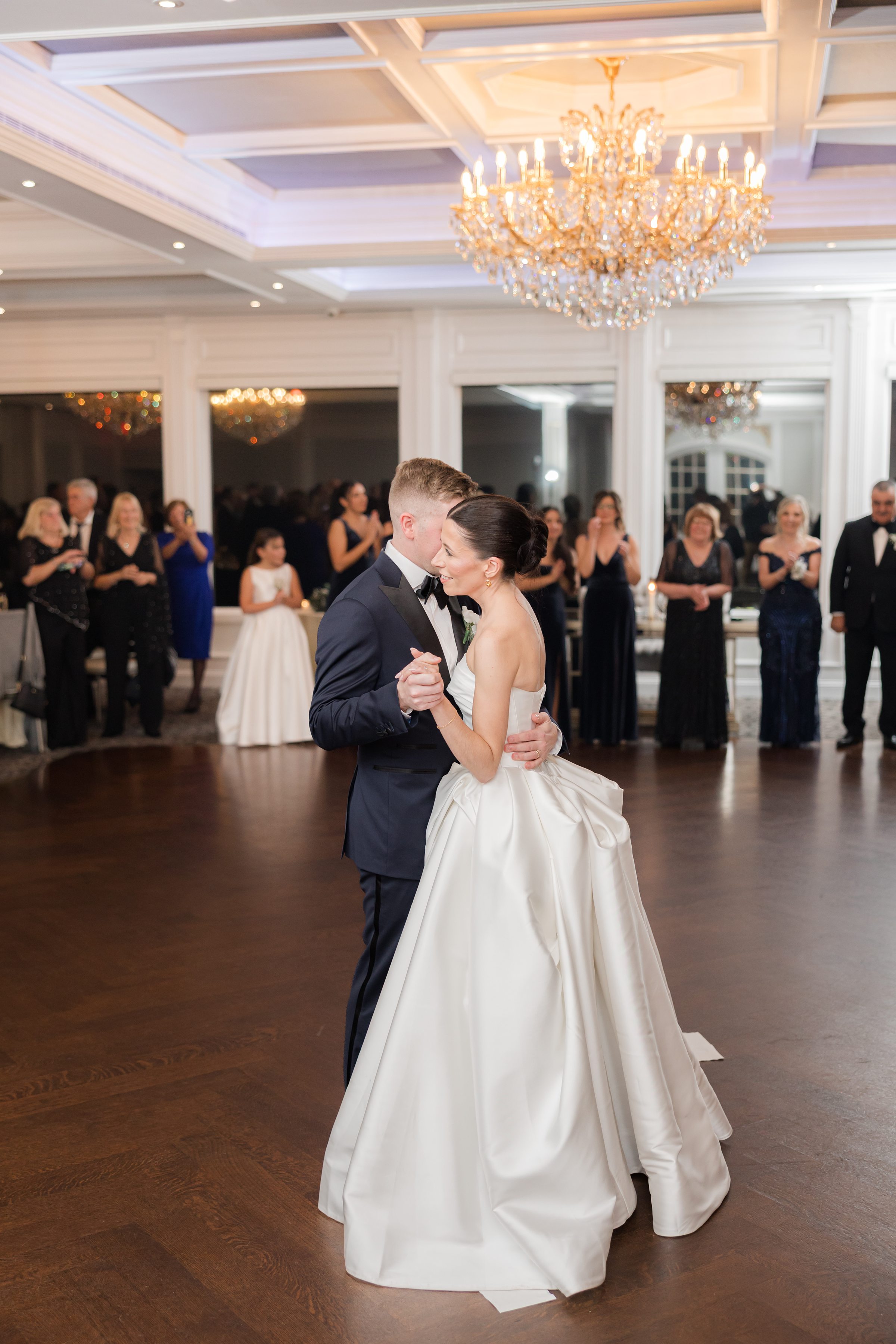 Bride and groom dancing, holding each other closely under chandelier light.