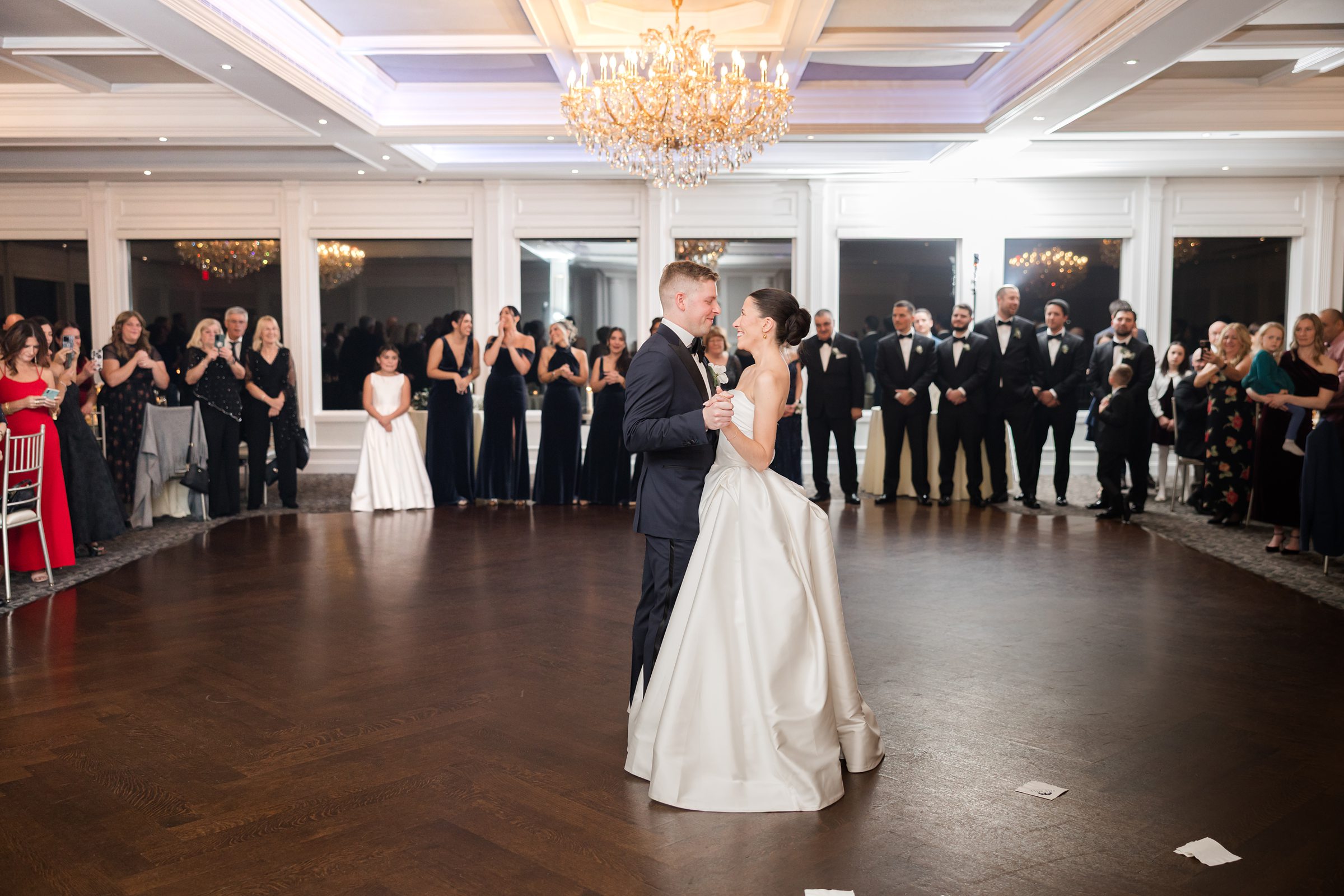 Bride and groom sharing their first dance in a bright ballroom under a large chandelier, surrounded by guests watching.