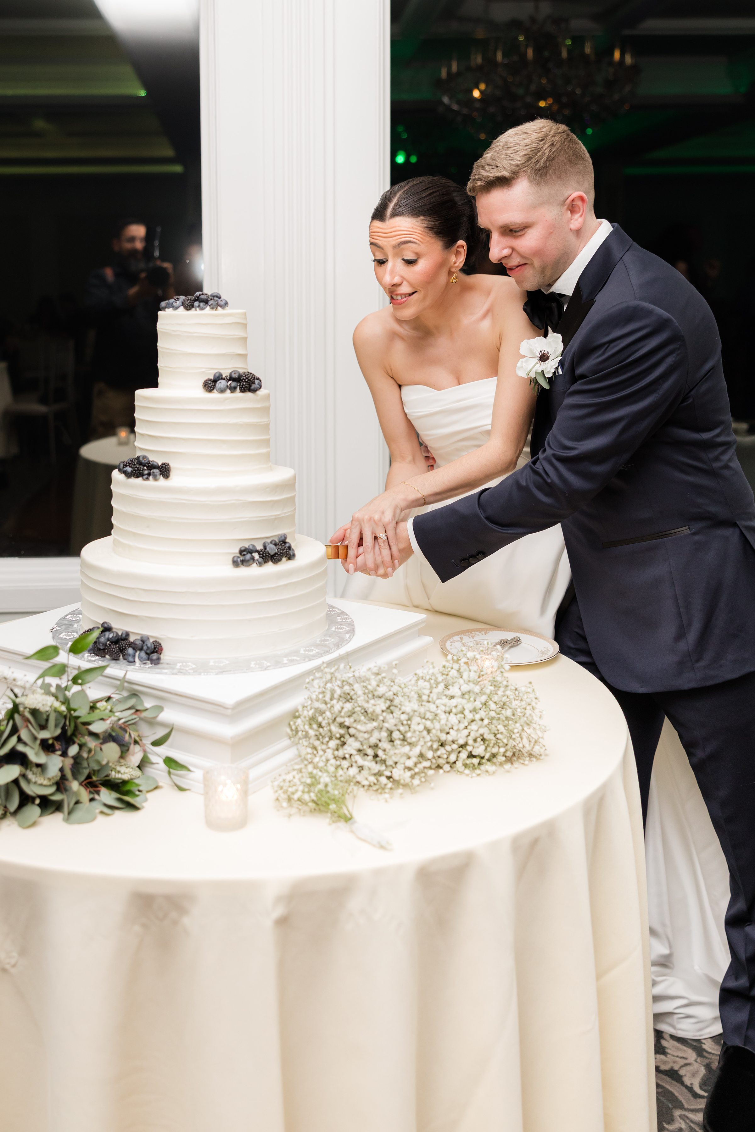 Bride and groom cutting a multi-tier white wedding cake decorated with berries, smiling together at the reception table.