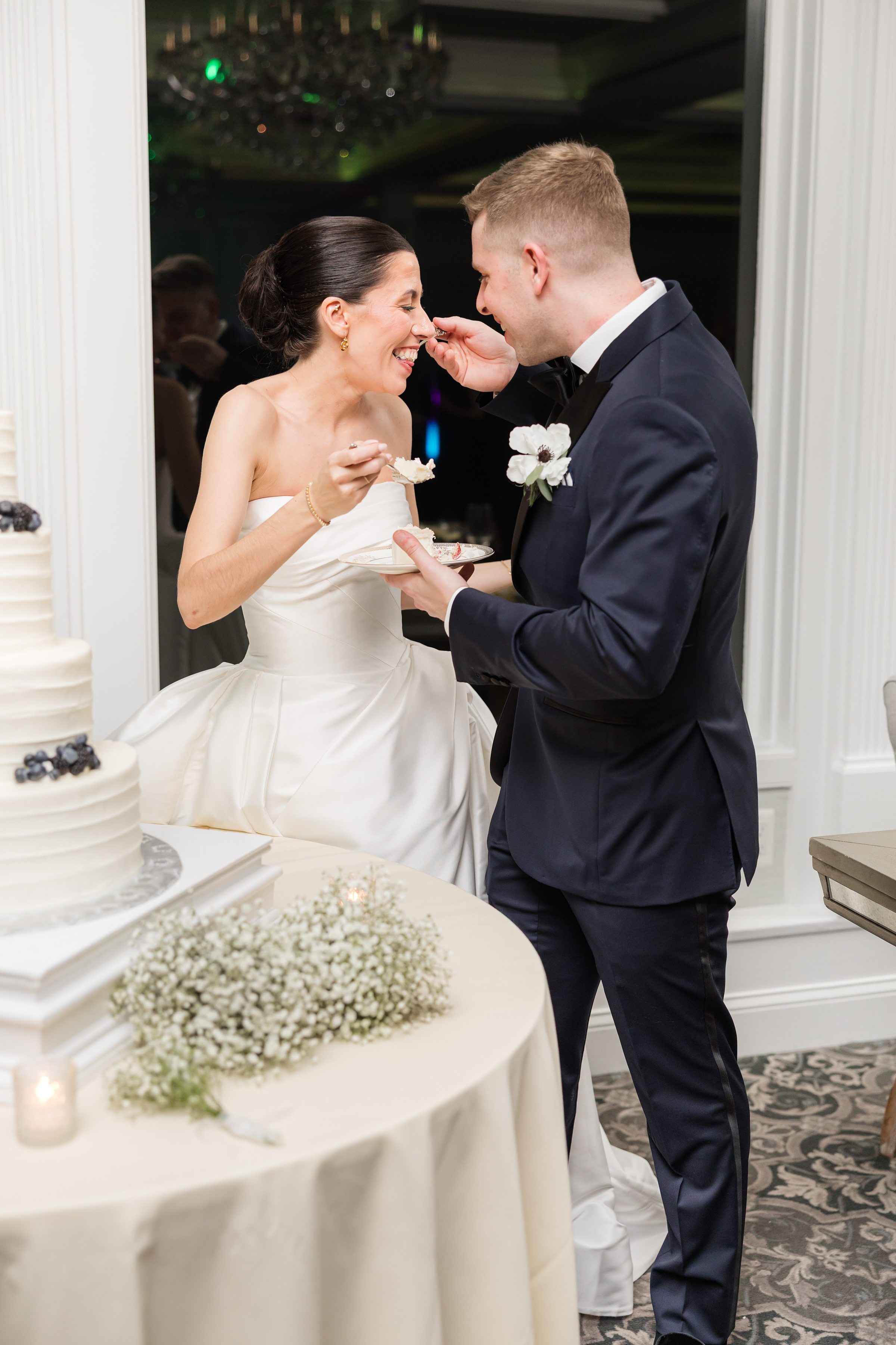 Bride and groom laughing while feeding each other cake beside a multi-tier white wedding cake.