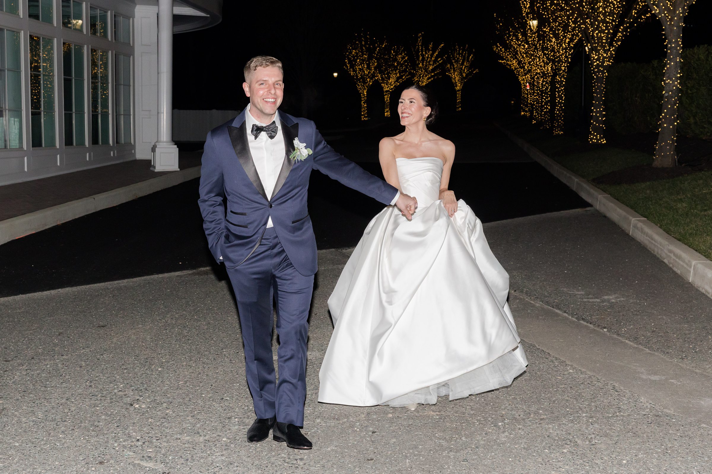Newlyweds walk together down a lit driveway at night, smiling at each other, with glowing trees lining the path.
