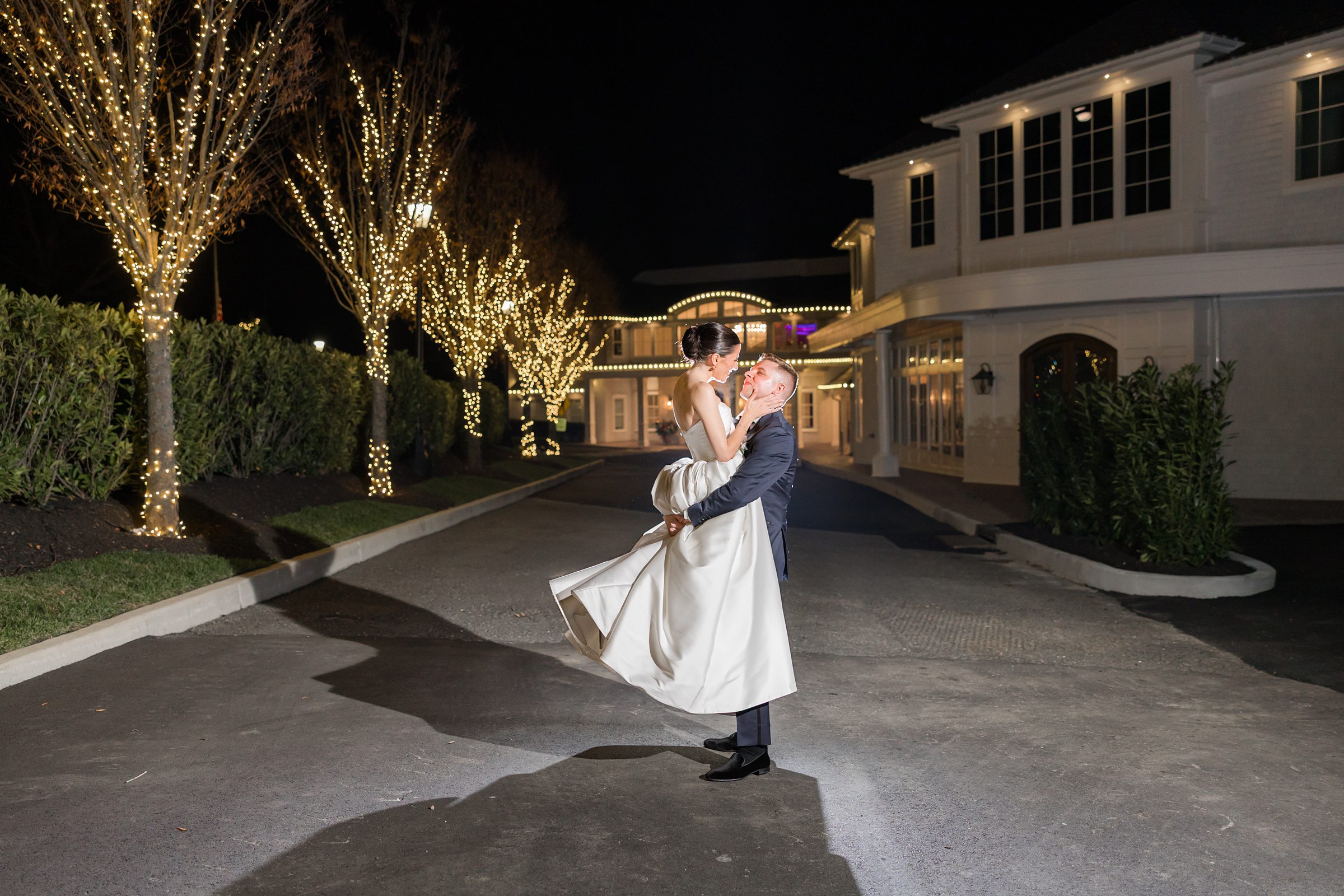 Bride and groom embrace outside at night, surrounded by trees wrapped in warm white lights, as he lifts her in front of a large, elegant venue.