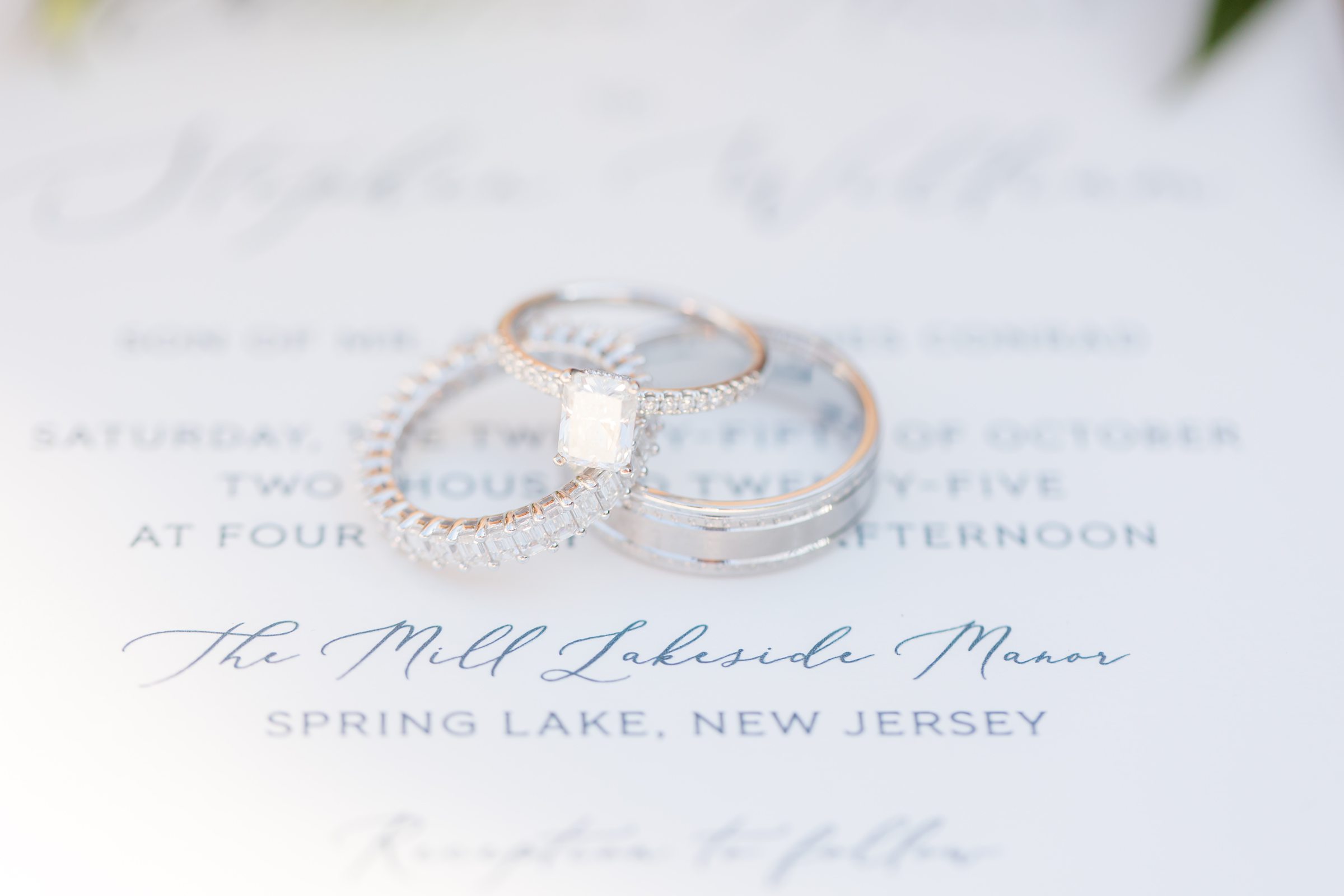 Engagement ring and wedding bands resting on a wedding invitation with visible venue text.