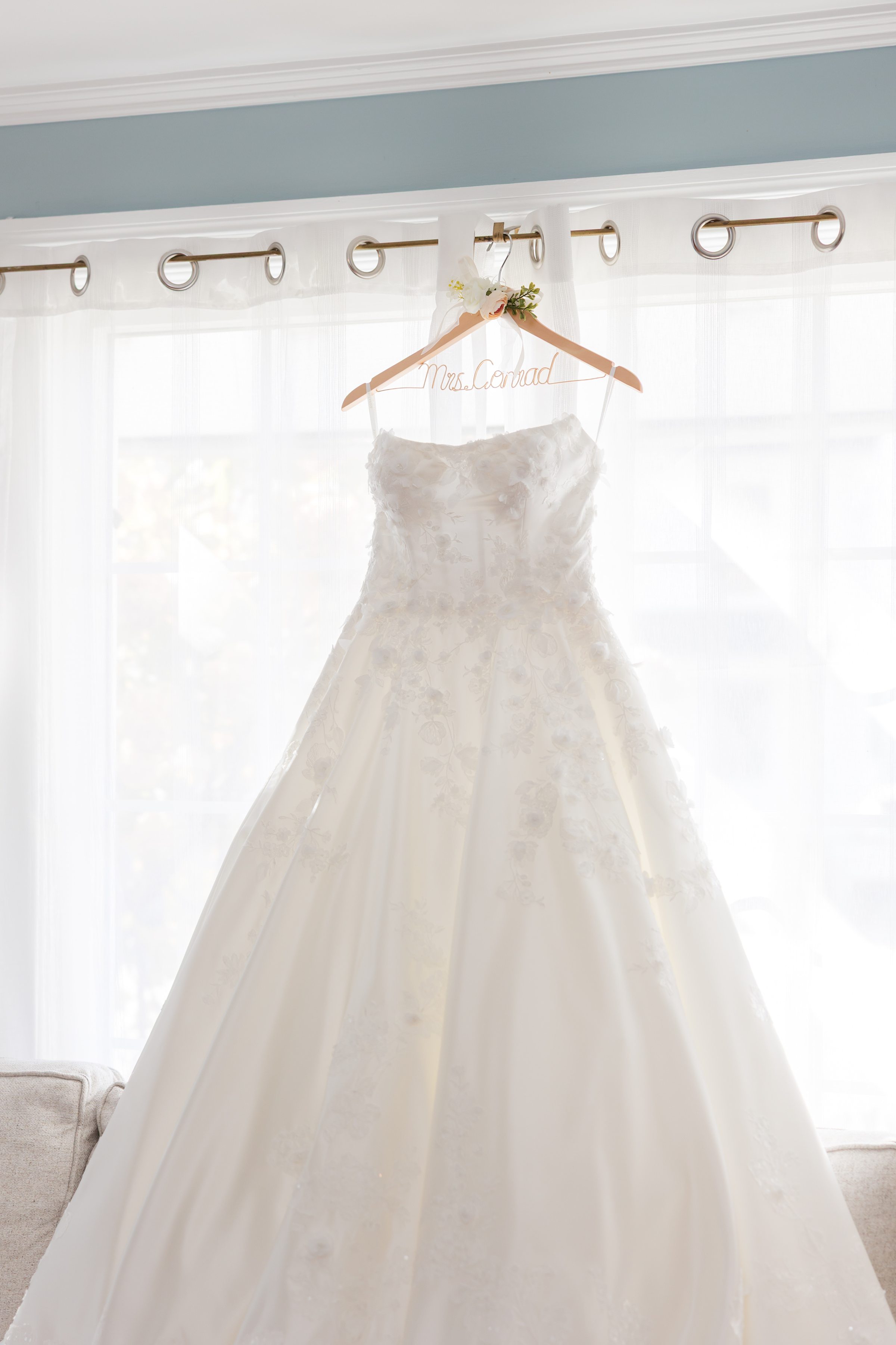 Wedding dress hangs on a personalized hanger in front of sheer white curtains with soft daylight behind.