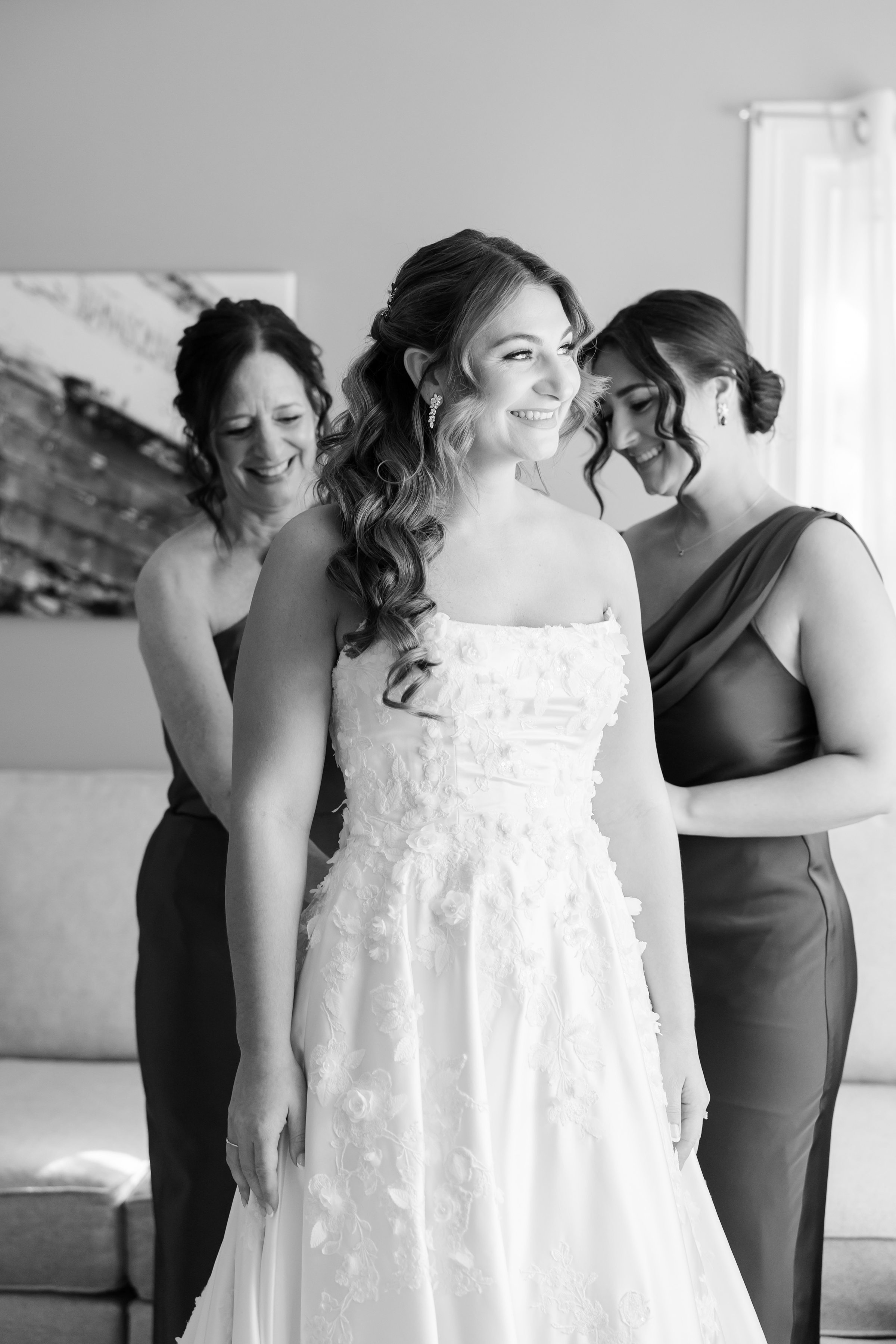 Bride smiling as her mother and sister stand behind her, lovingly helping adjust her wedding dress in a bright room.