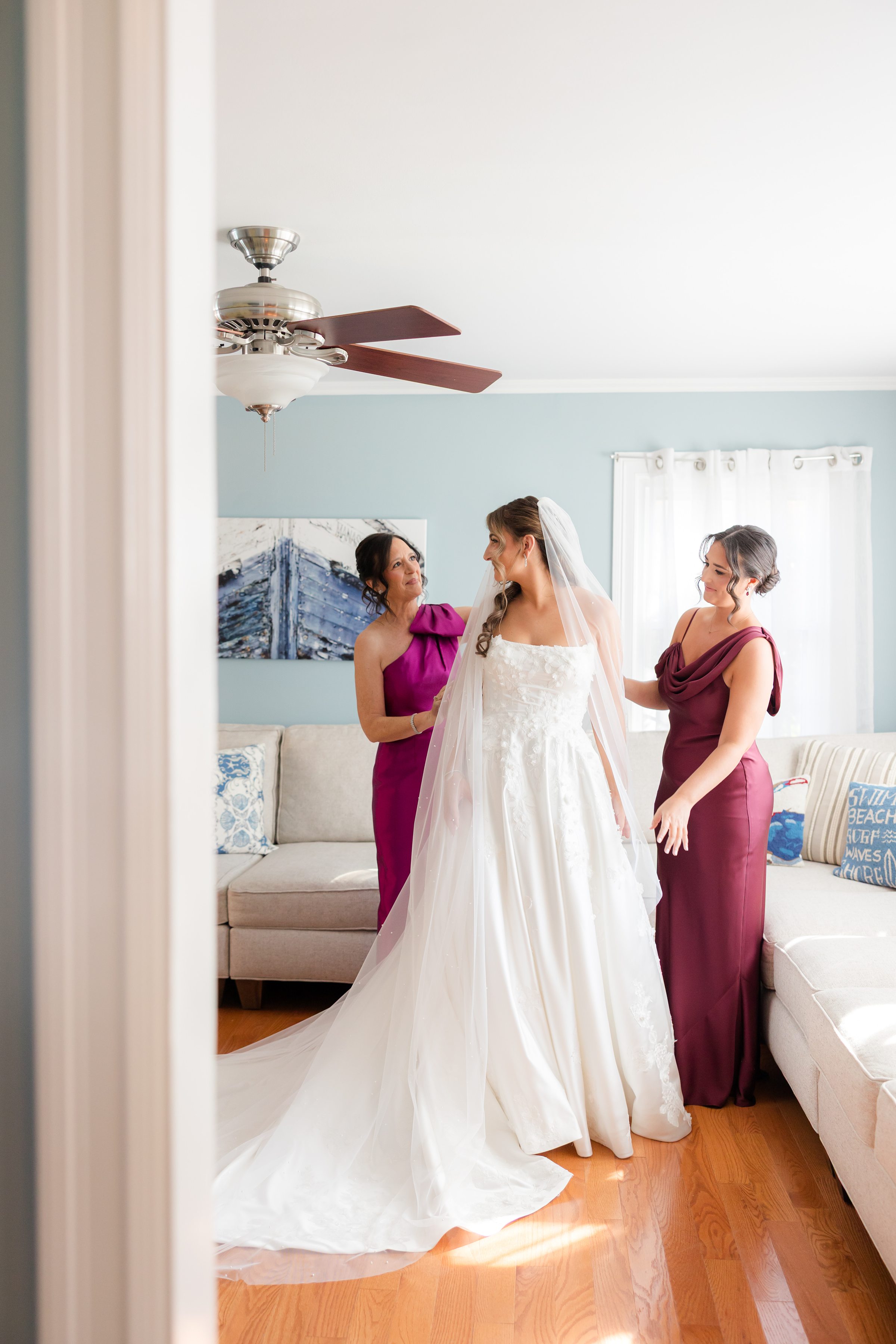 Bride in her wedding gown shares a warm moment with her mother and sister as they stand together, smiling and adjusting her dress in a softly lit room.
