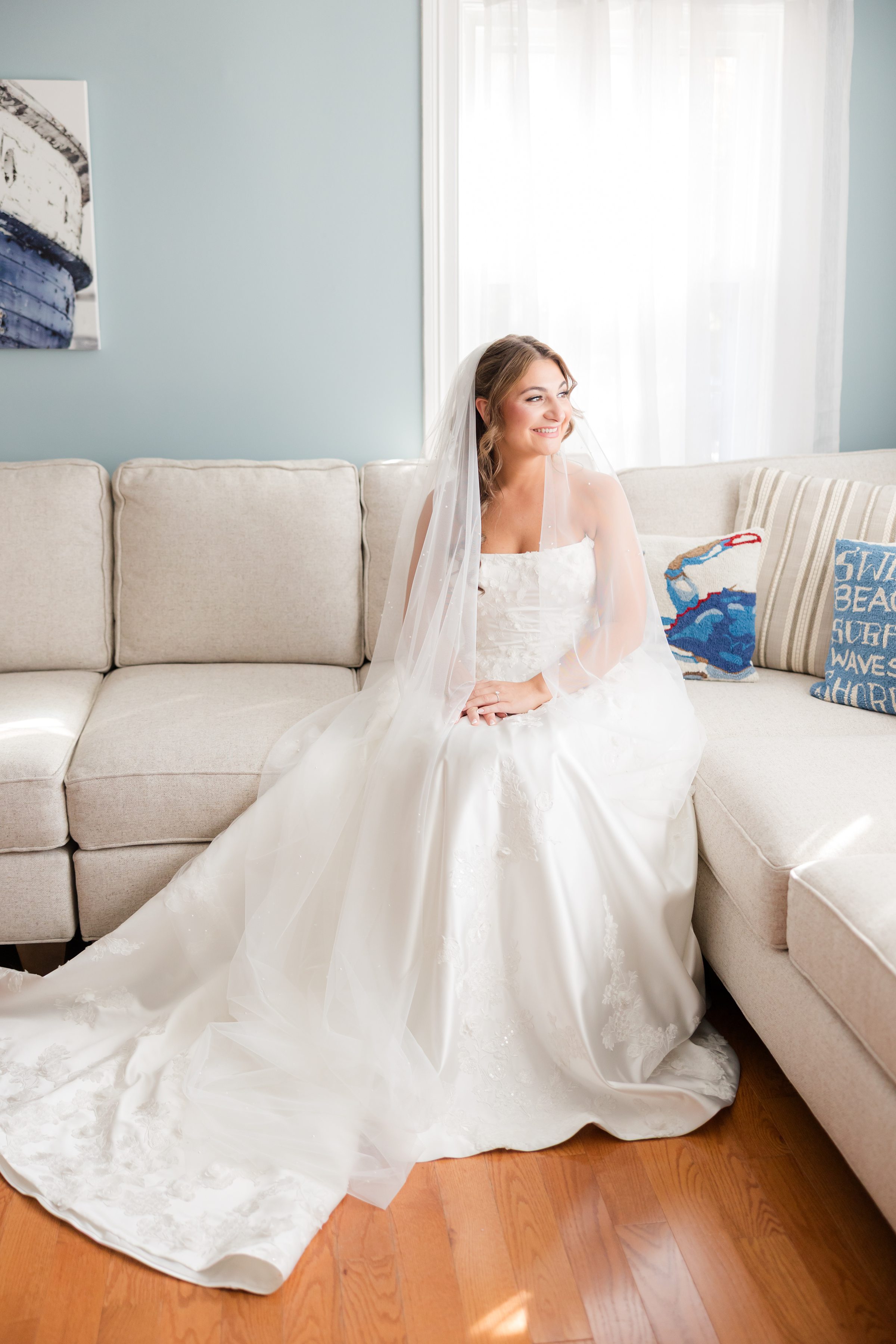 Bride sits on a beige sectional sofa in a sunlit room, her long veil and train spread across the wooden floor as she looks off to the side.
