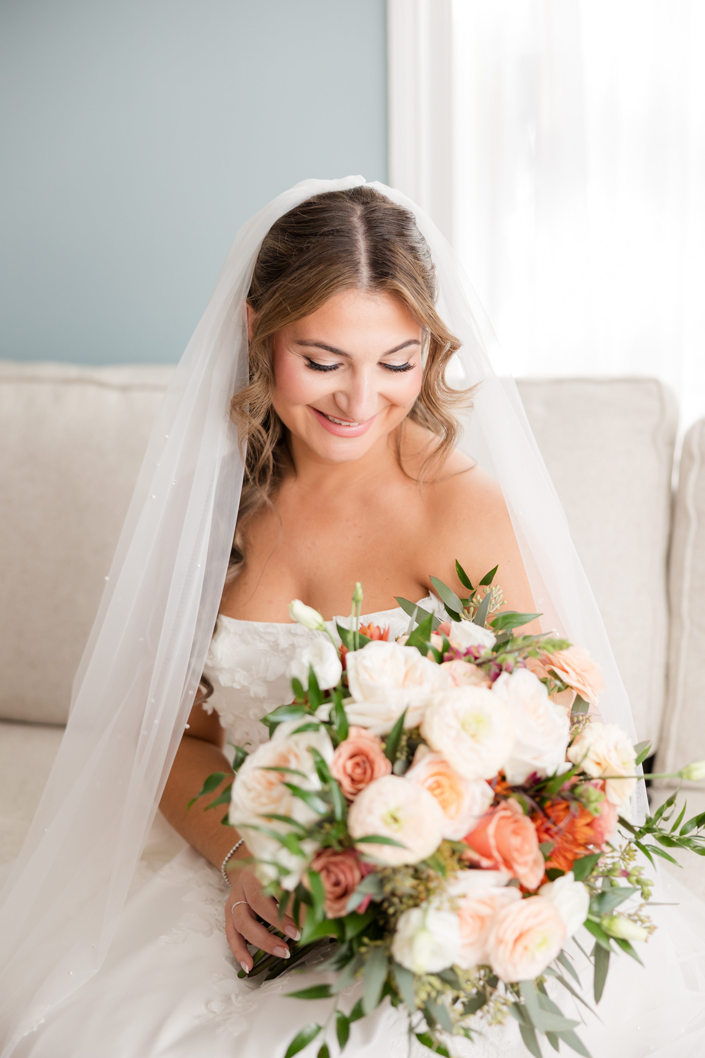 Bride in a strapless white wedding gown and veil smiles down at a bouquet of peach, white, and orange flowers while seated on a light-colored couch.